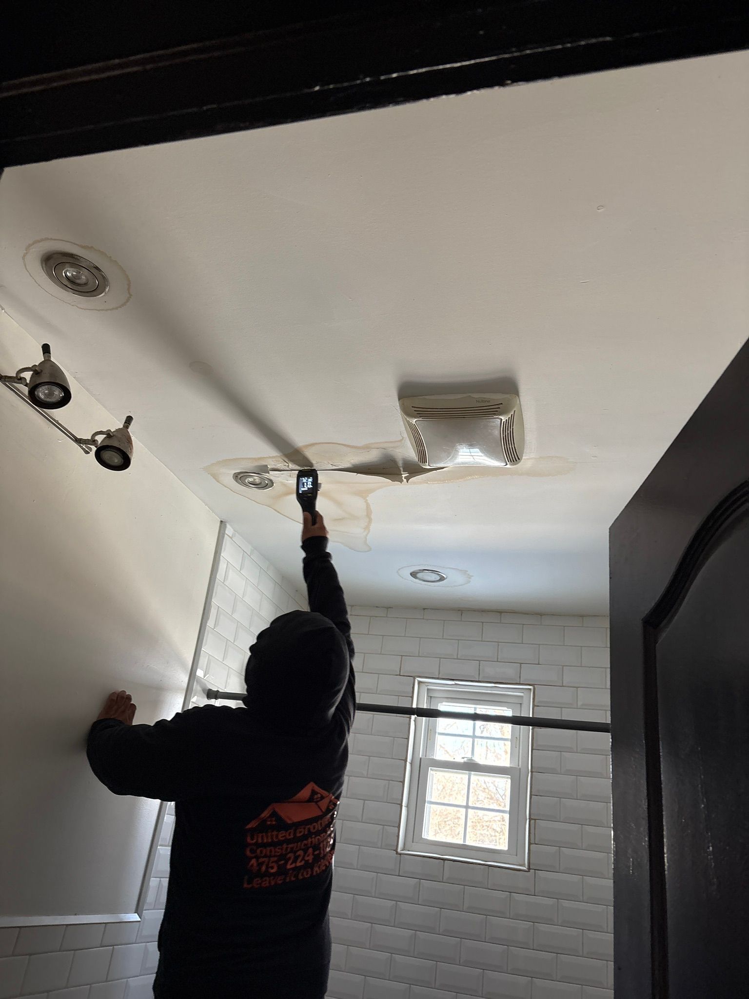 Person painting a ceiling around a vent in a white tiled bathroom using a roller and extension pole