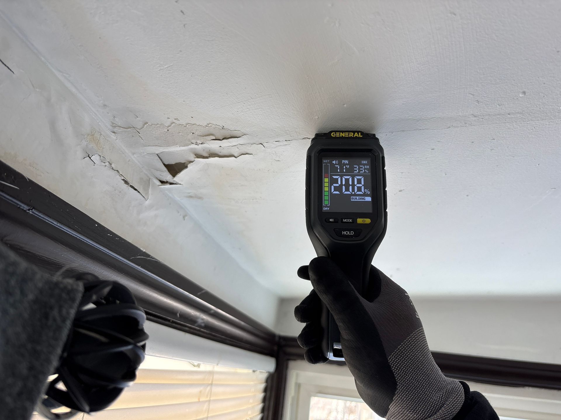 Gloved hand holds a thermal camera up to a damaged white ceiling near exposed pipes.