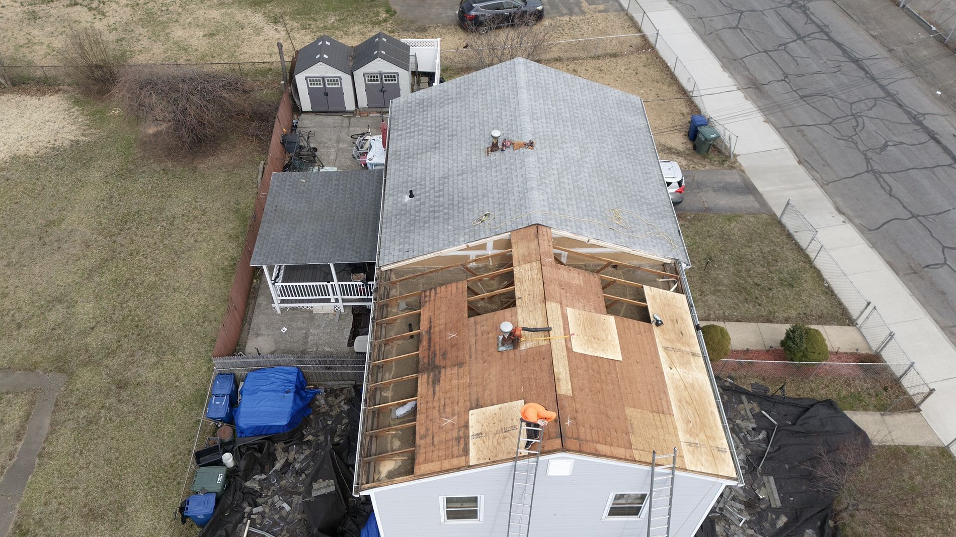 Aerial view of a house with a damaged roof, neighboring sheds, and a driveway beside a street.