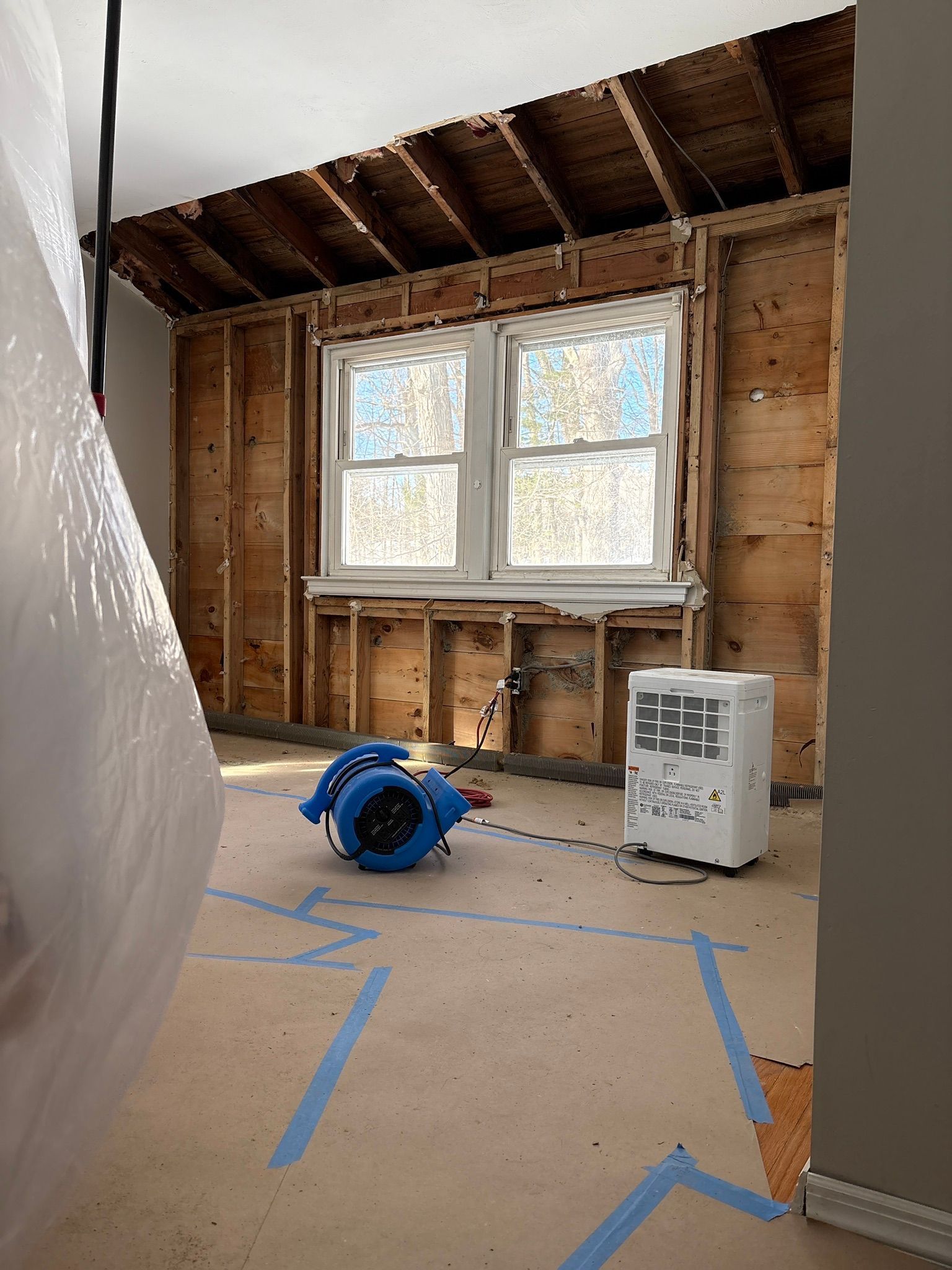 Partially renovated room with exposed studs, two windows, taped floor, a fan, and a portable heater