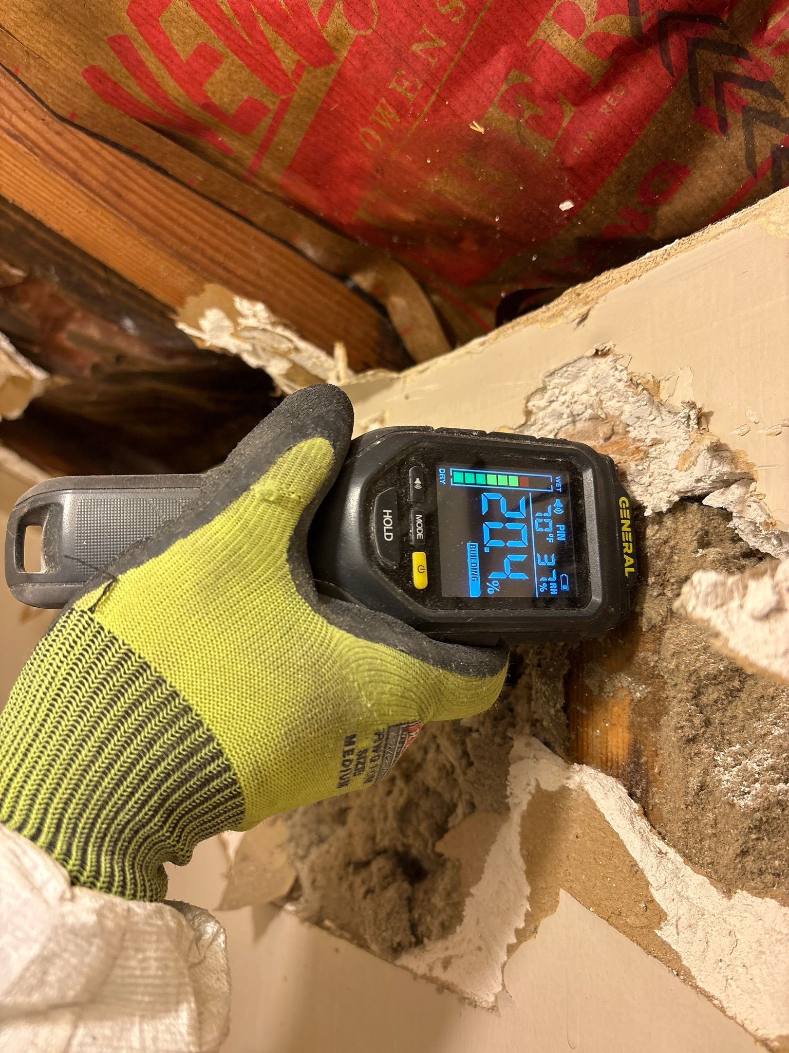 Gloved hand holds a digital moisture meter against a damaged wall, reading 22.4.