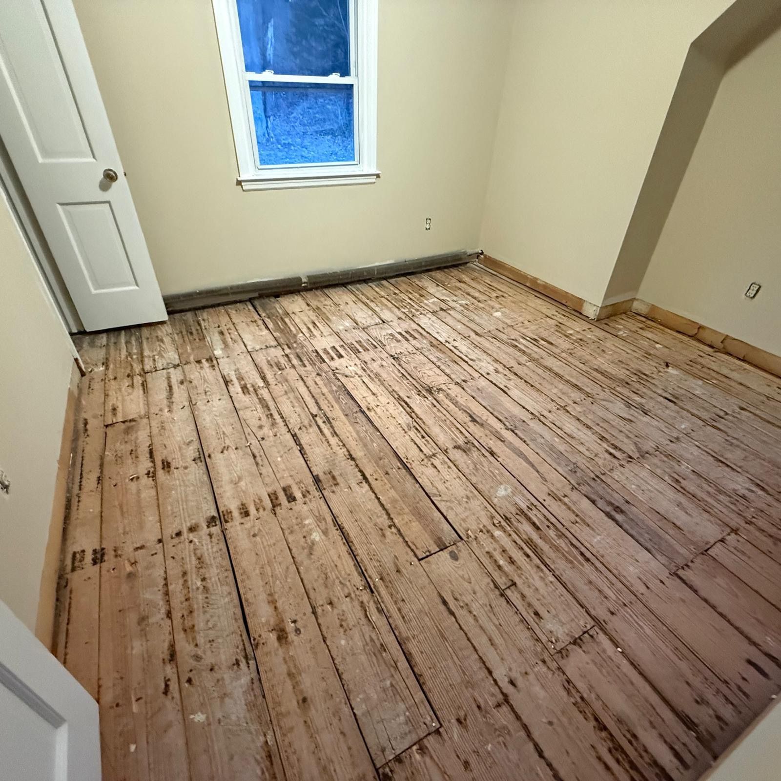 Empty room with unfinished wooden floor, cream walls, and a small window.