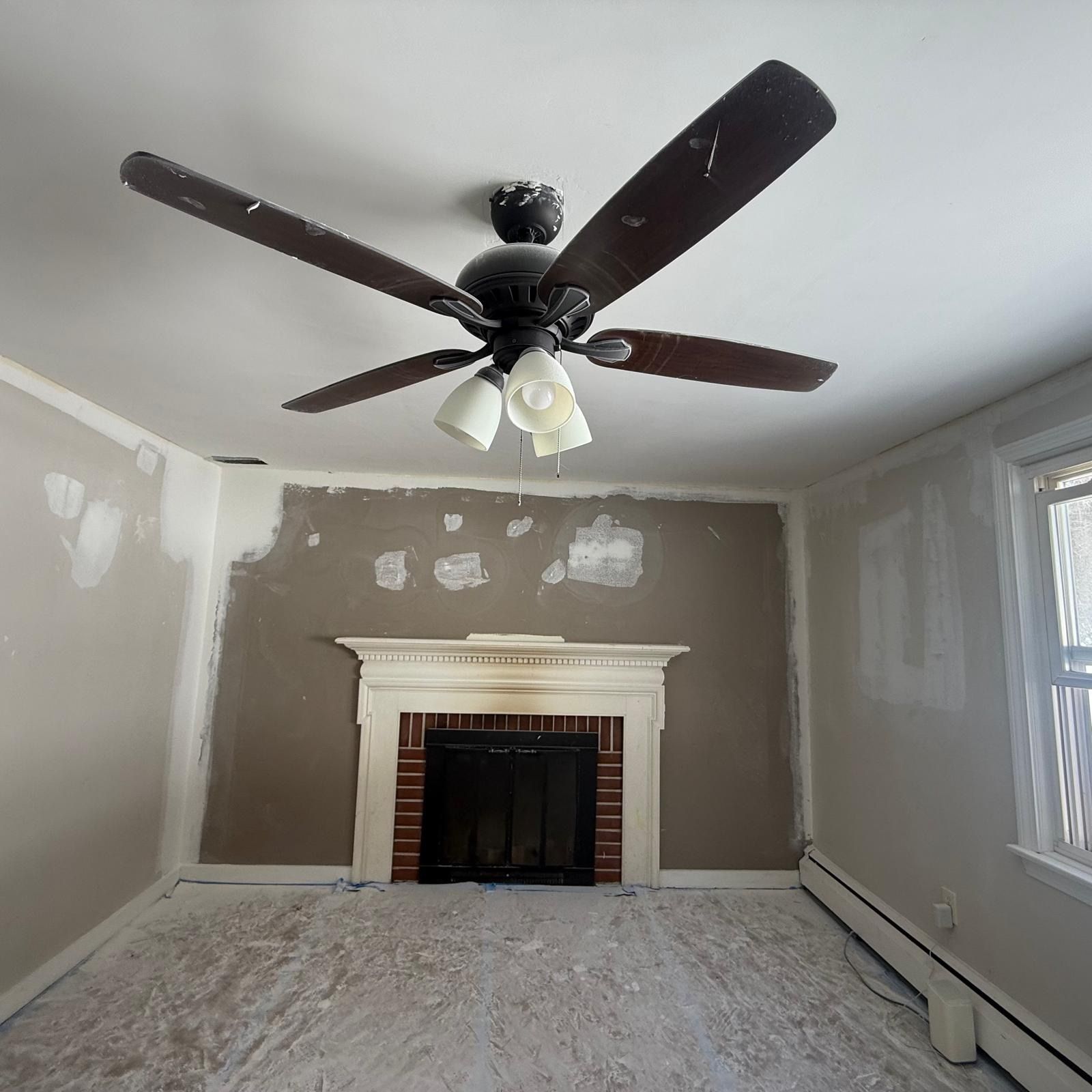 Empty beige room with fireplace, dark ceiling fan, and large windows during renovation