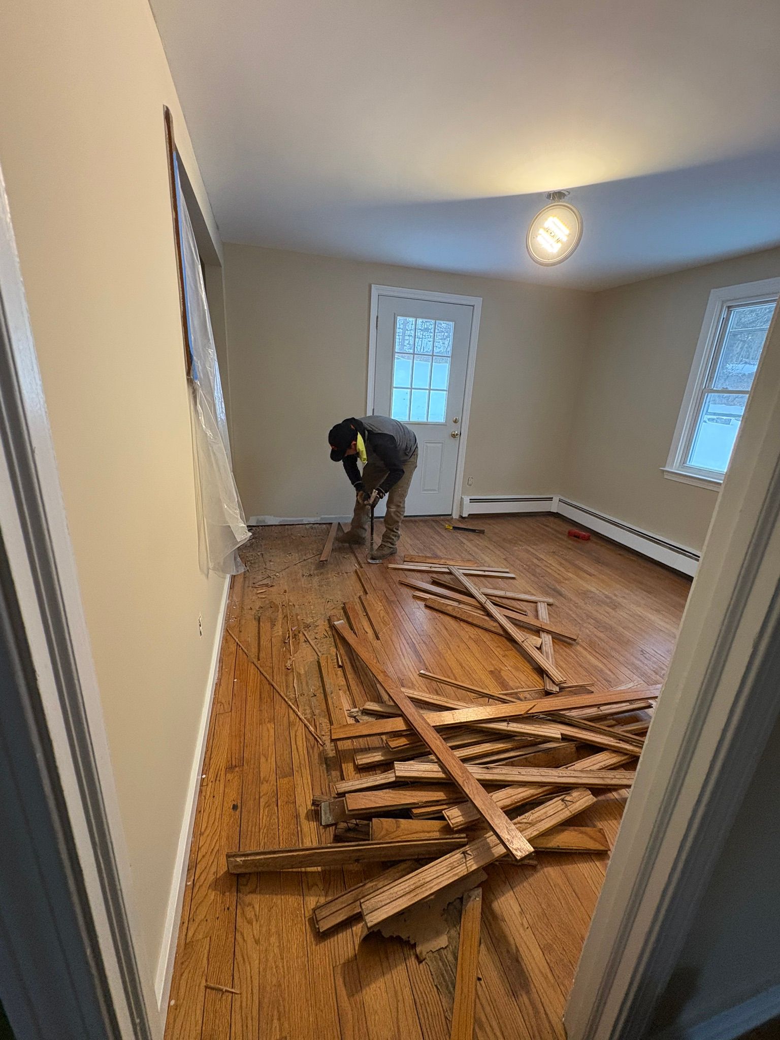 Worker removing broken hardwood flooring in an empty room with a window and ceiling light.