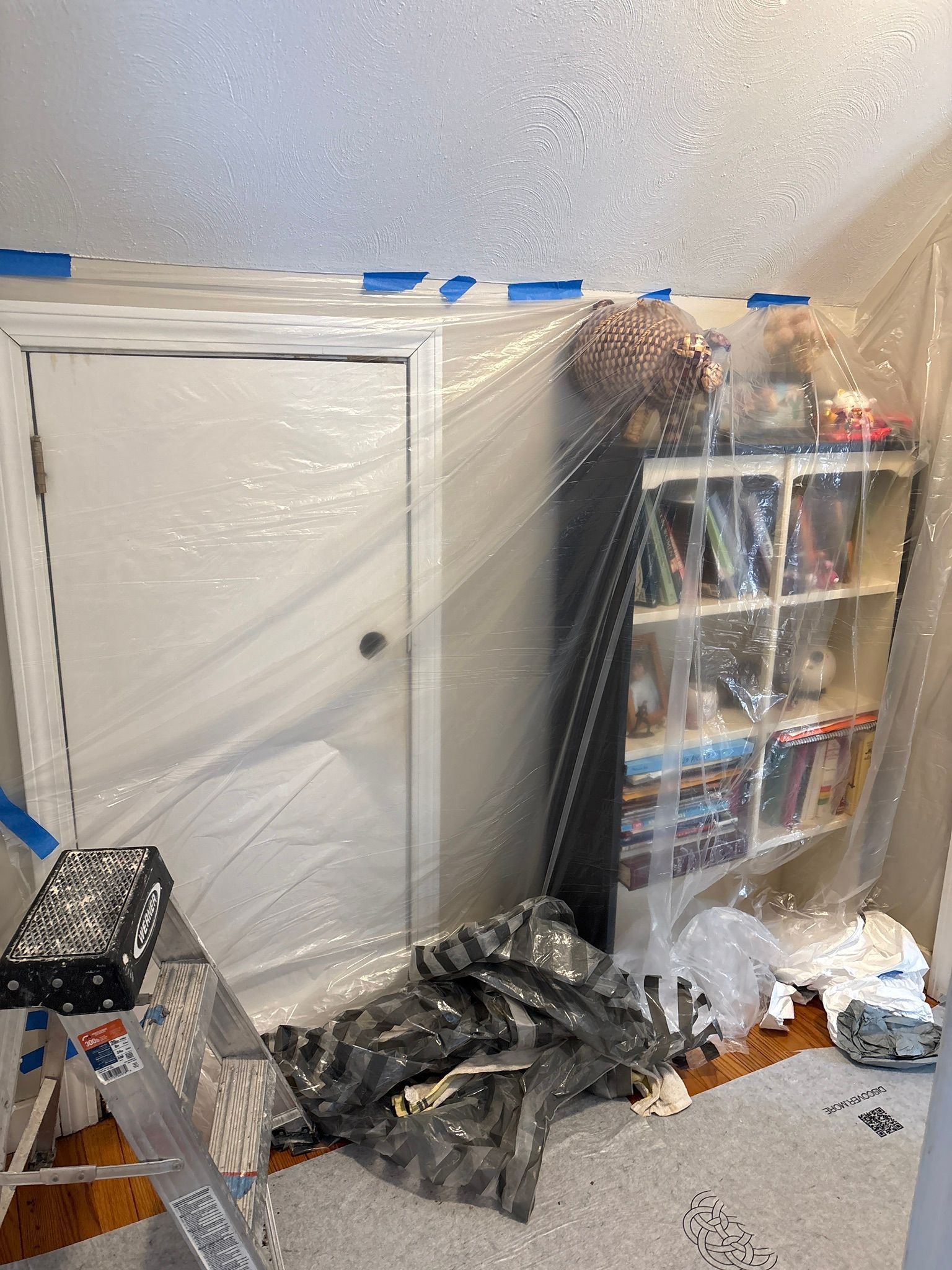 Cluttered room with plastic-covered door, shelving unit, and bags piled on the floor.