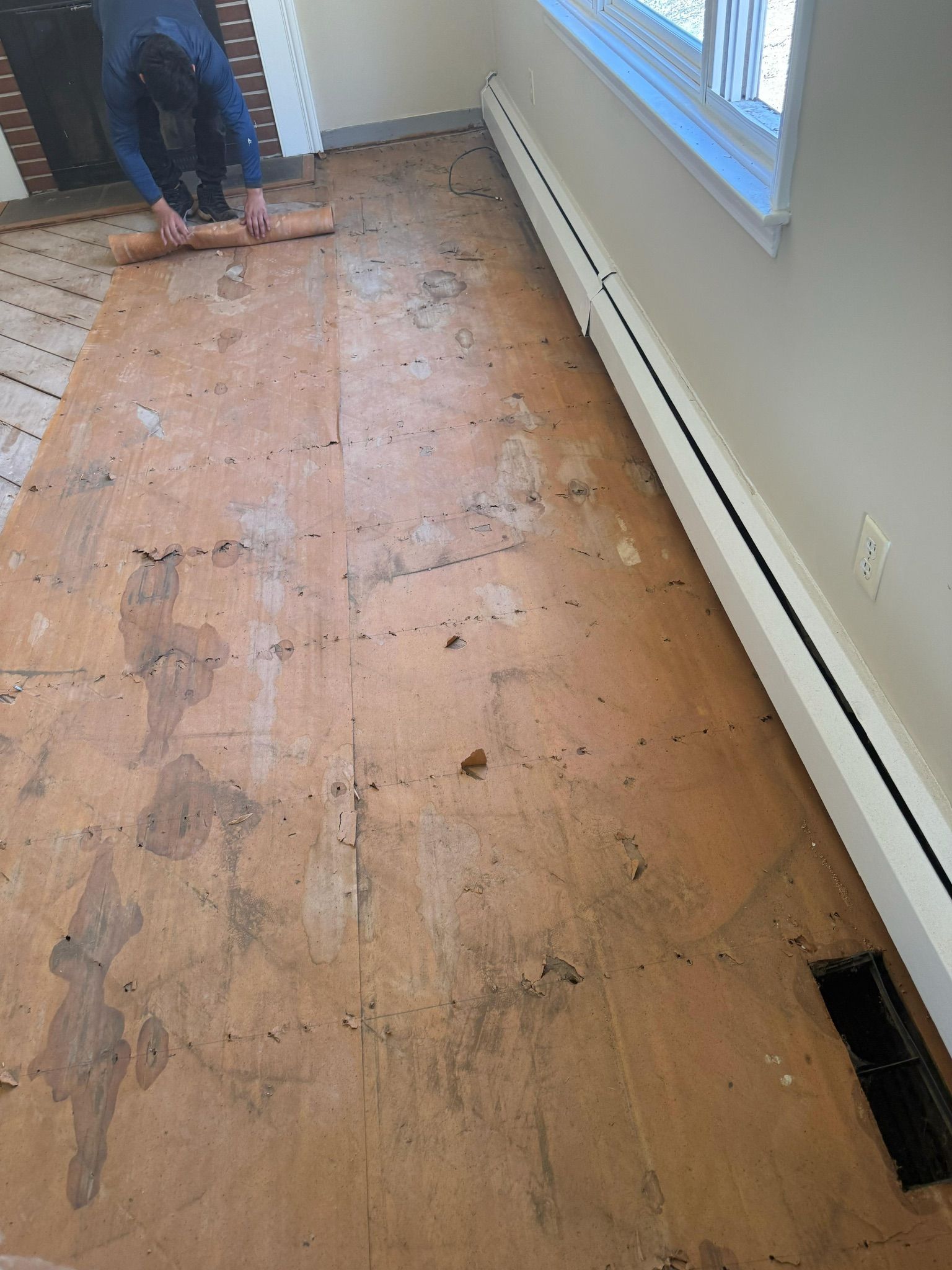 Person kneeling on a bare plywood floor in a room, working near a wall and window.