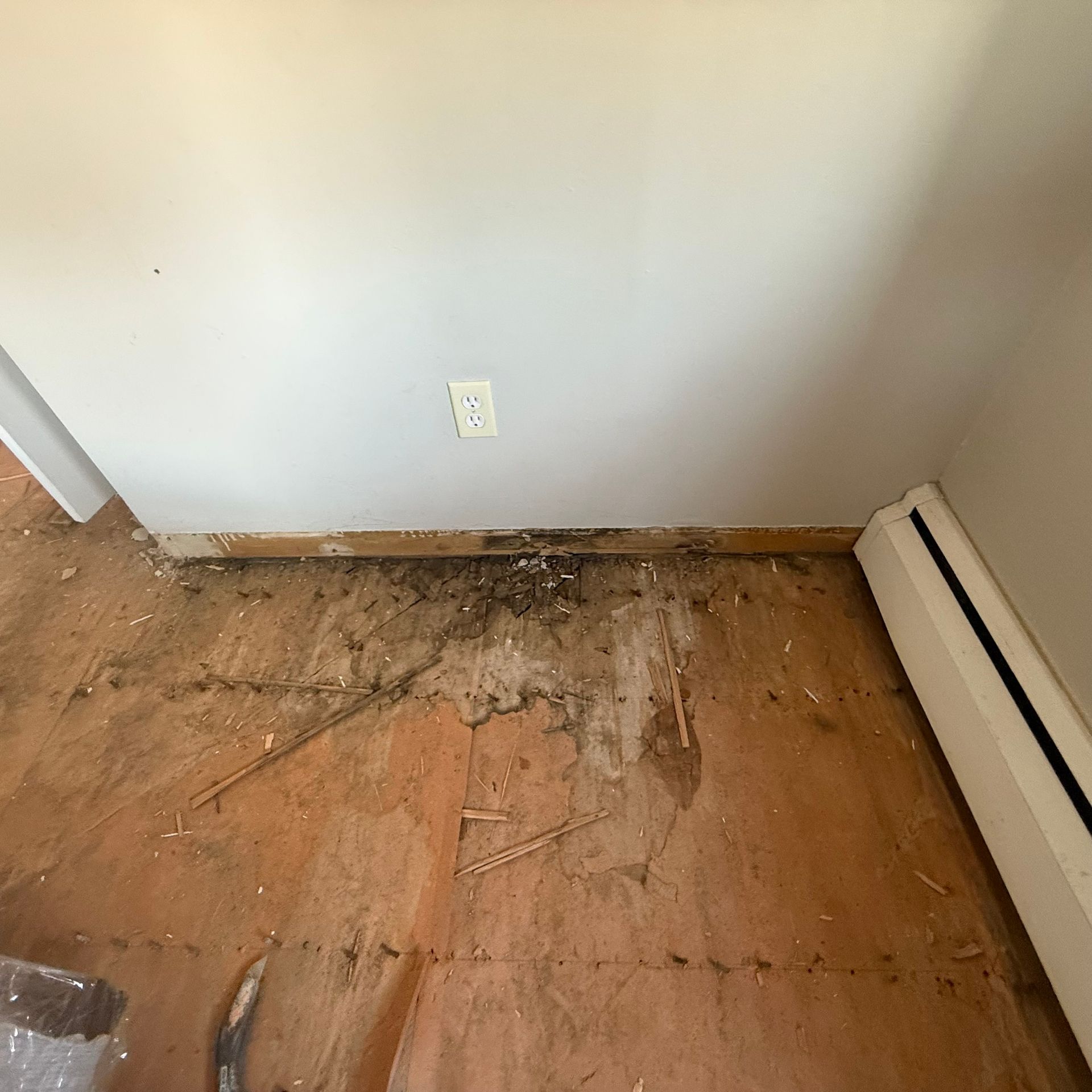Damaged floor with water stains and rotting wood along a wall near an outlet and baseboard heater