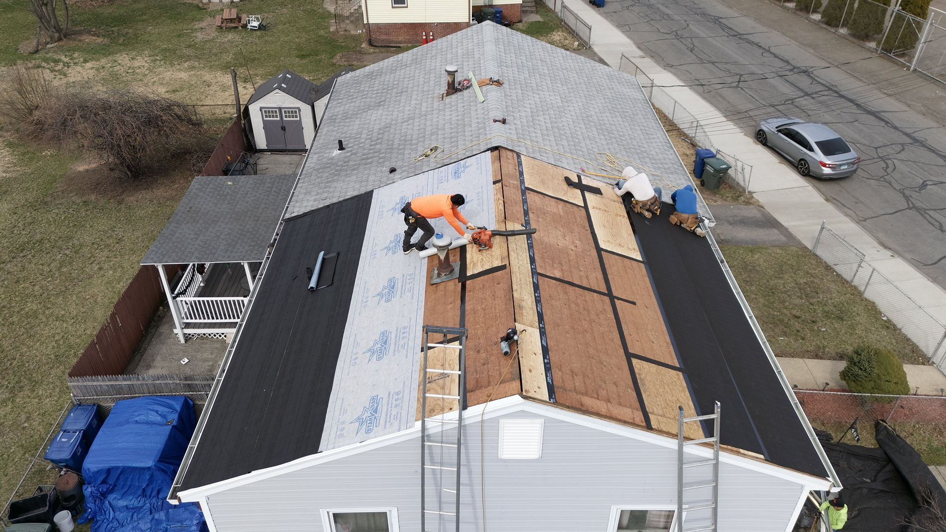 Roofers installing shingles on a house roof beside a street, with materials and tools spread across the roof.