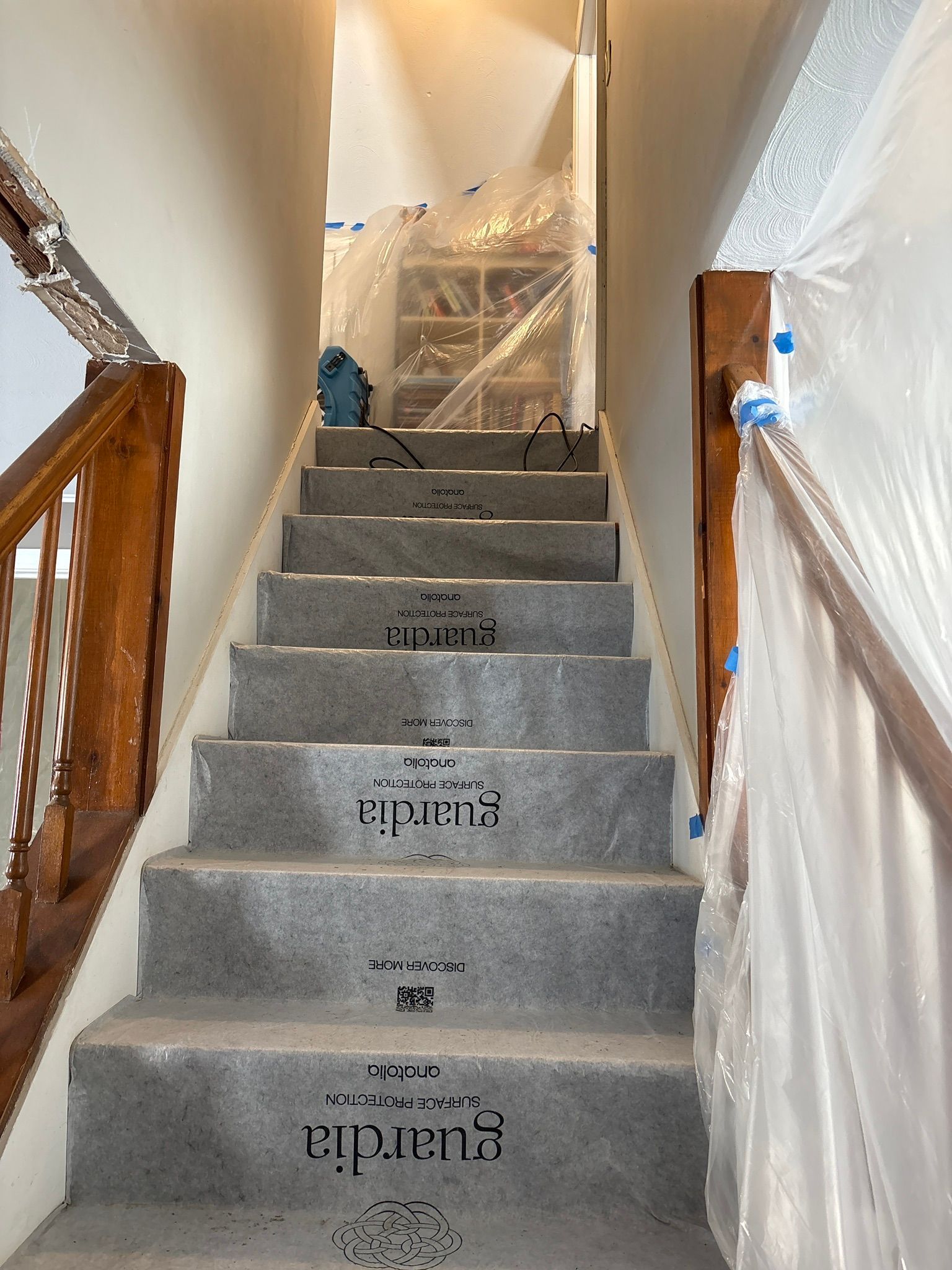 Carpeted staircase under renovation, with plastic sheeting and a wooden handrail on both sides.