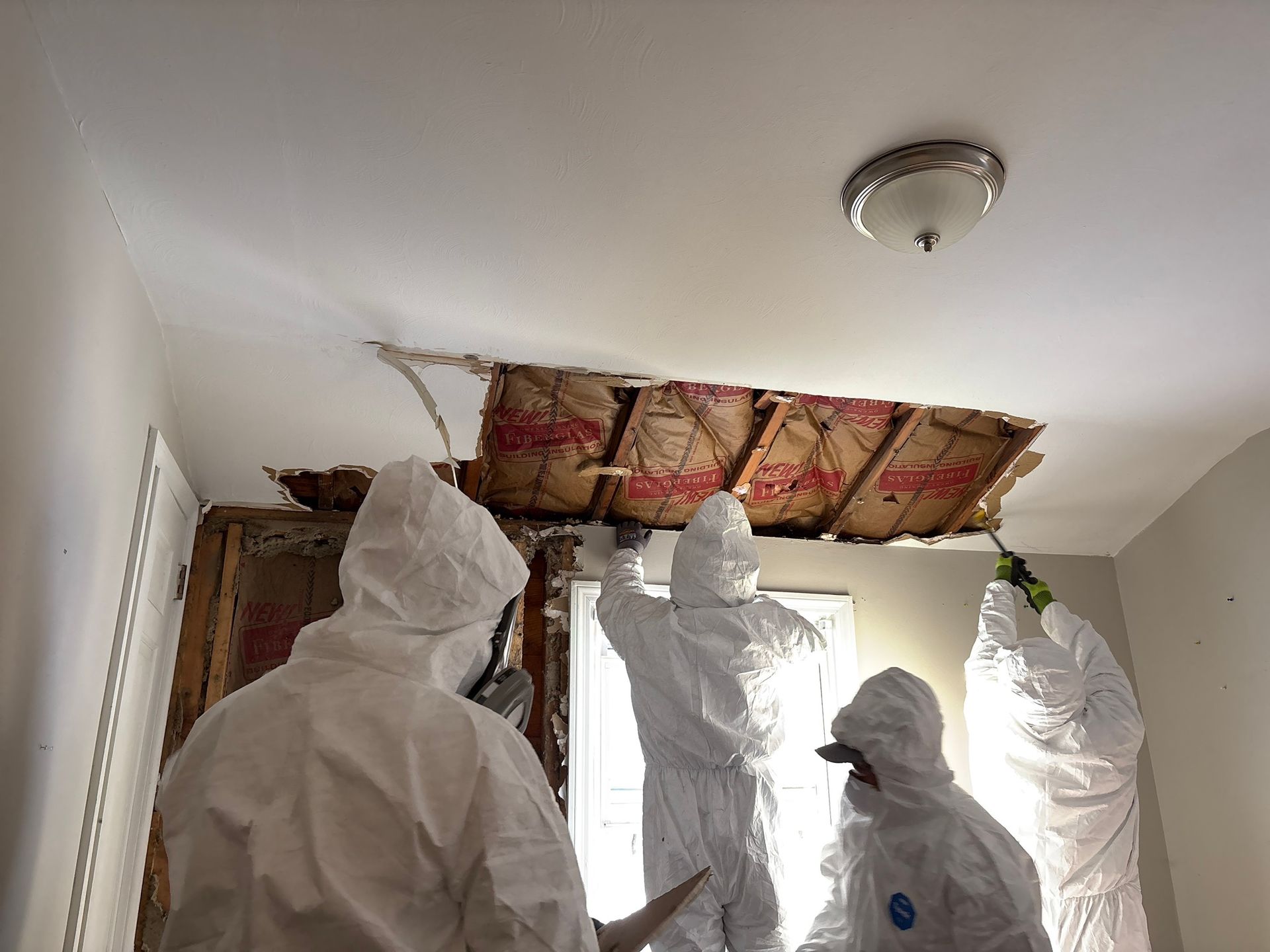 Three people in white protective suits inspecting exposed insulation in a ceiling repair area.