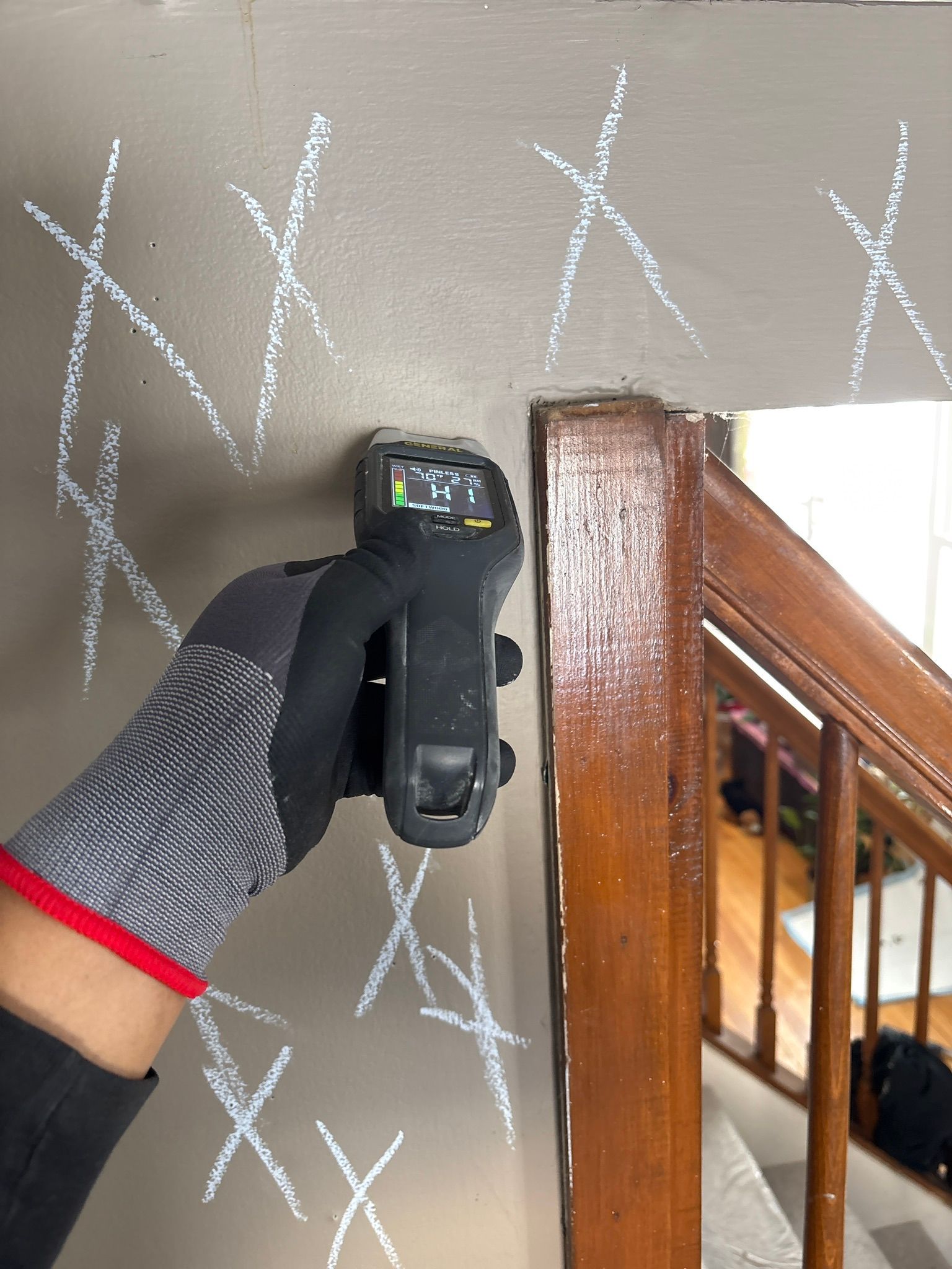 Gloved hand holding a black handheld device against a gray patterned wall beside a wooden stair rail