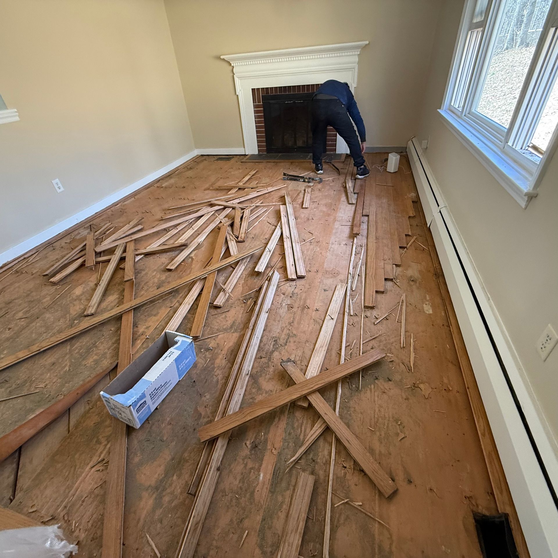 Person clearing debris in a damaged room with torn hardwood flooring and a fireplace.