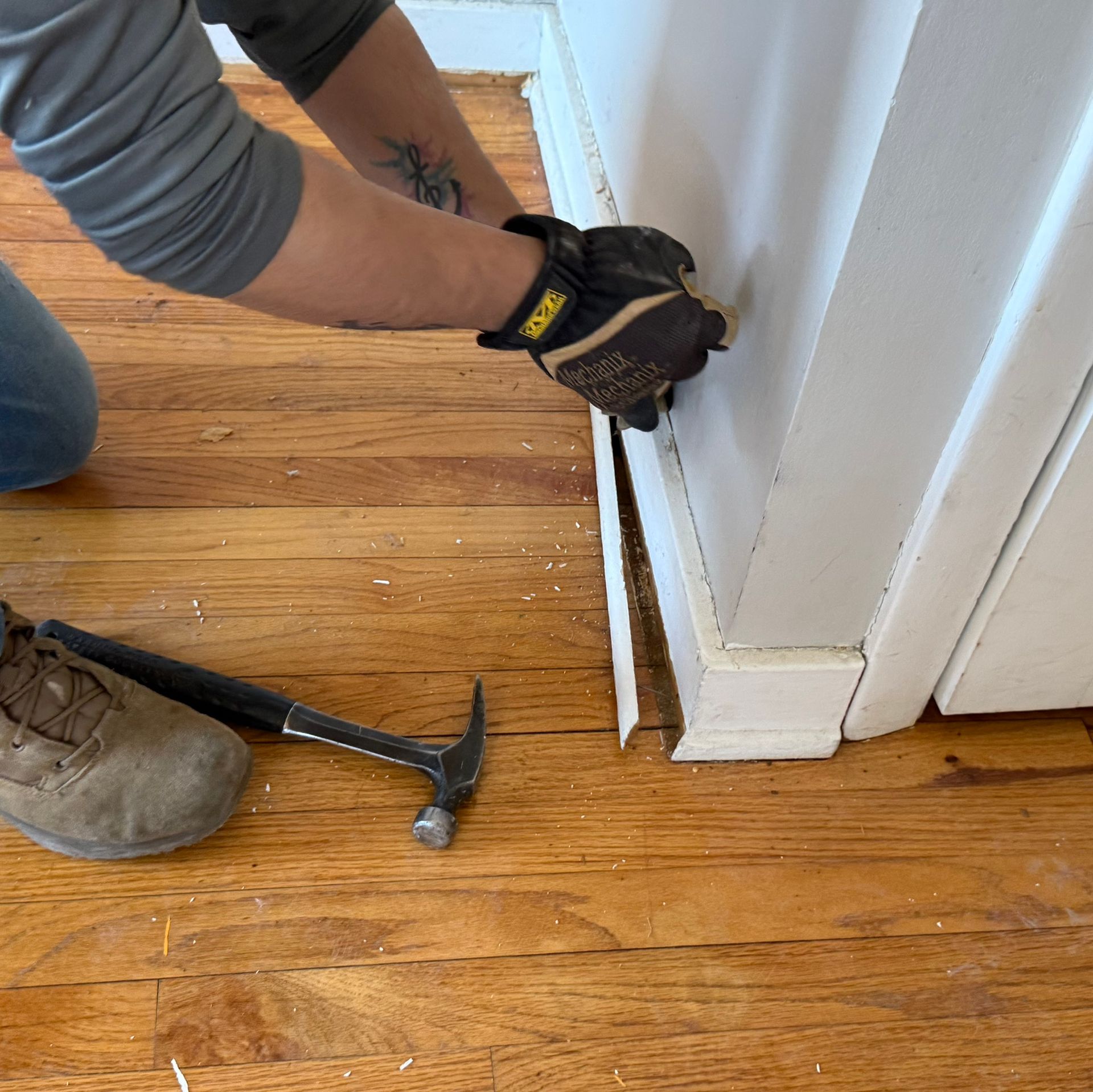 Person uses a pry bar and hammer to remove damaged trim beside a white wall on a hardwood floor.