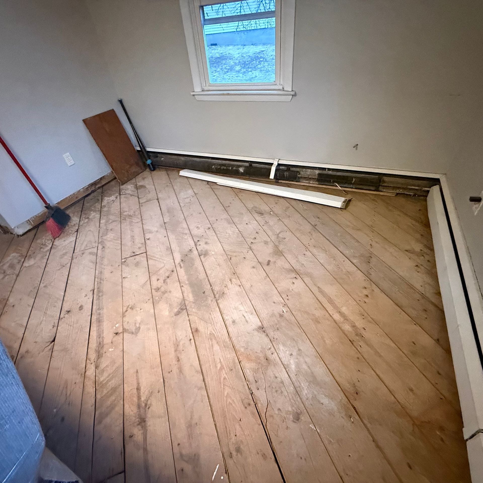 Empty room with bare wooden floorboards, a small window, and trim pieces leaning along the walls.