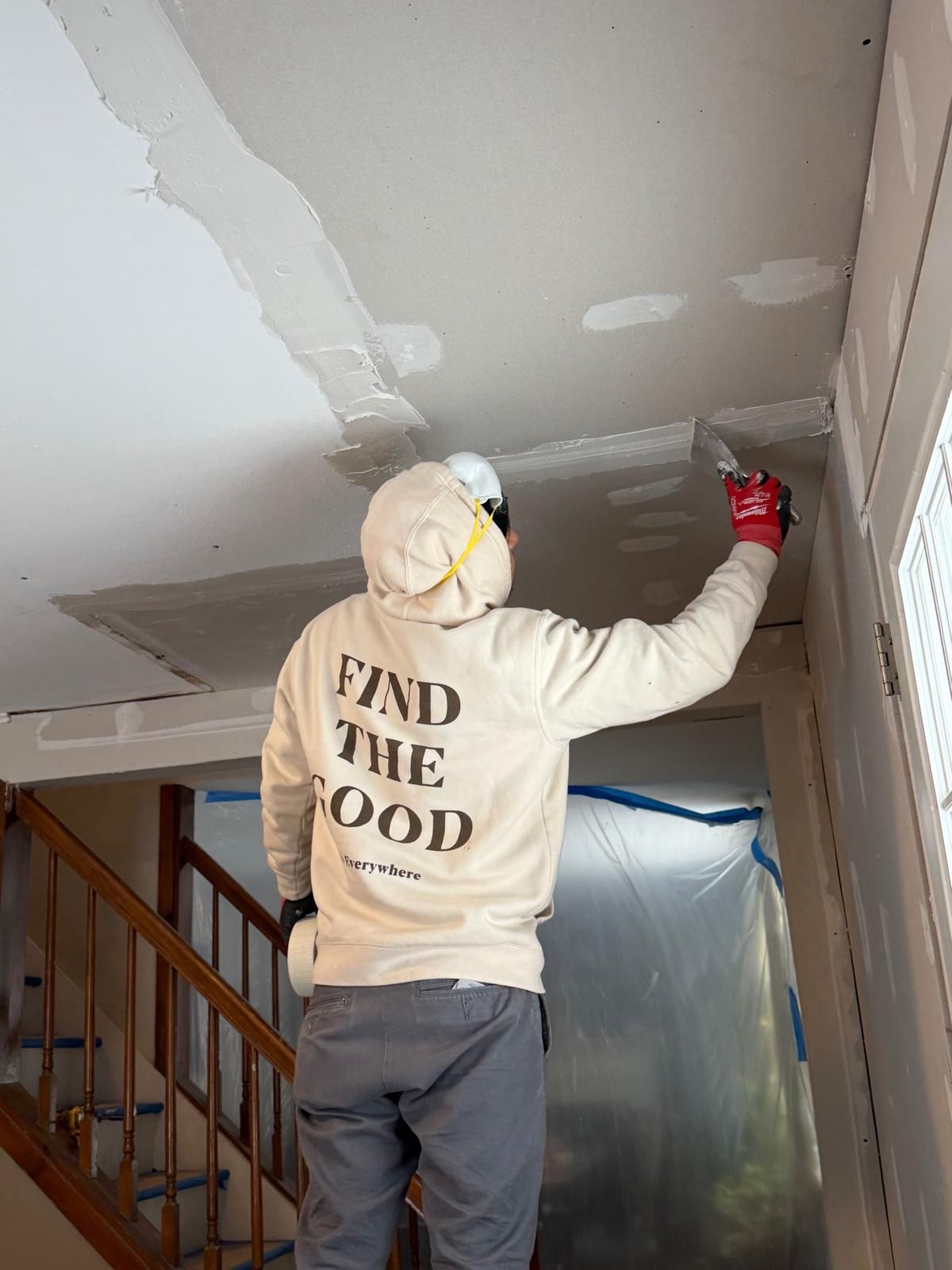 Person plastering and smoothing a damaged ceiling inside a stairwell, wearing a hoodie and gloves.