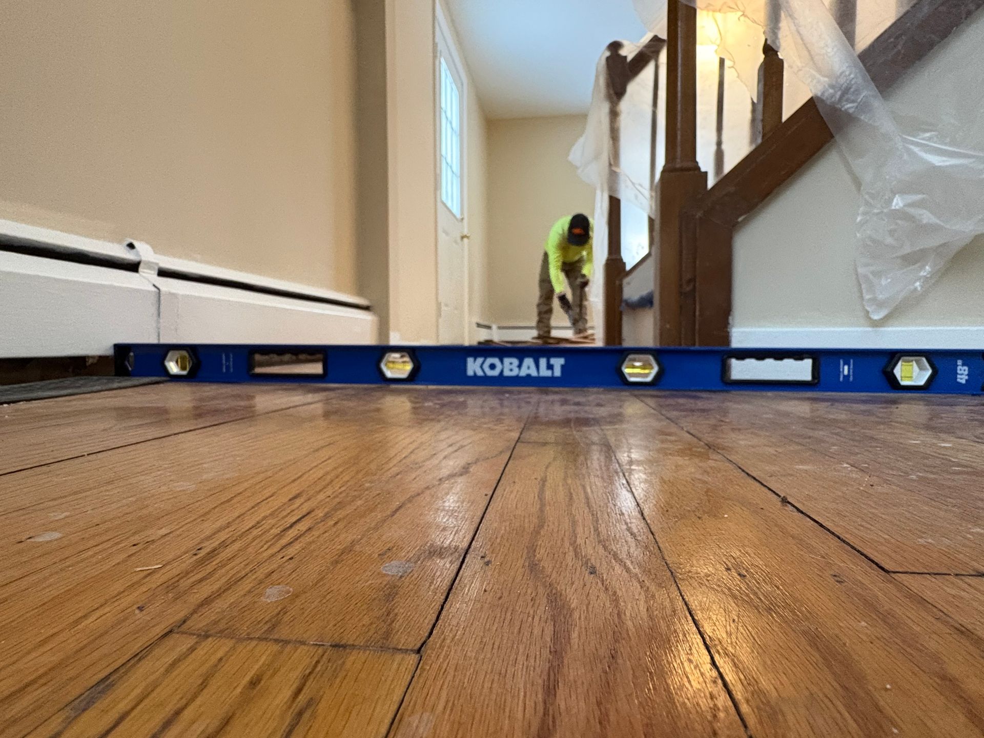 Low-angle view of a hardwood floor with a blue KOBALT level across the room near a staircase.