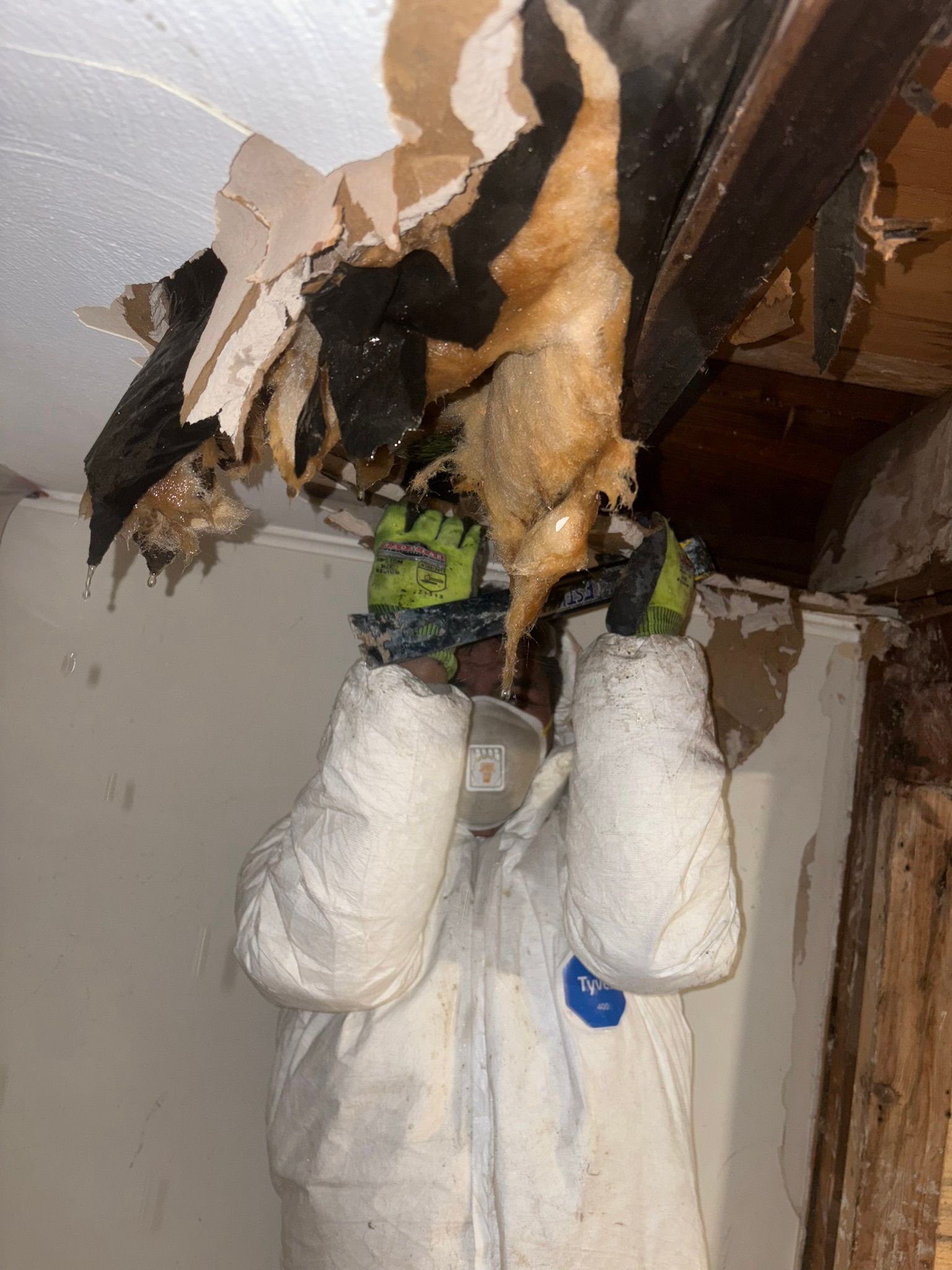 Worker in protective suit inspecting a damaged ceiling with exposed insulation and wooden beams