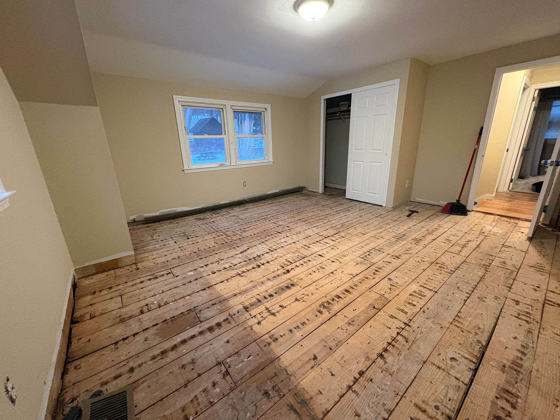 Empty beige room with unfinished wood floor, window, closet door, and overhead light