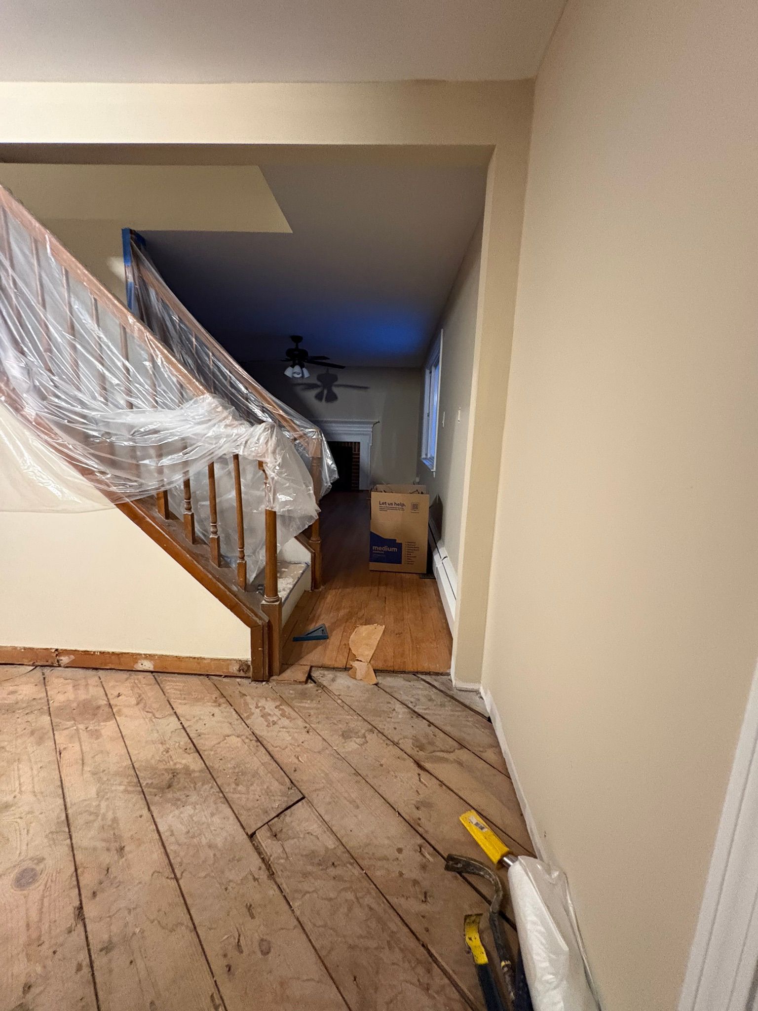 Narrow hallway with exposed wooden floorboards, staircase wrapped in plastic, and a blue-lit room in the distance