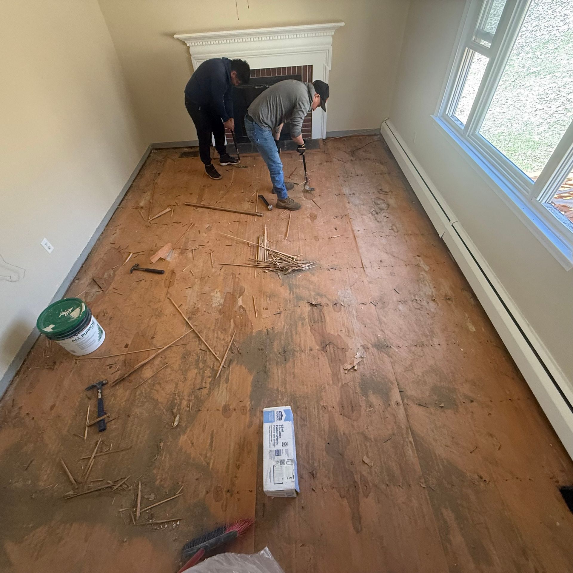 Two workers removing old flooring from a narrow room with windows and a fireplace mantel.