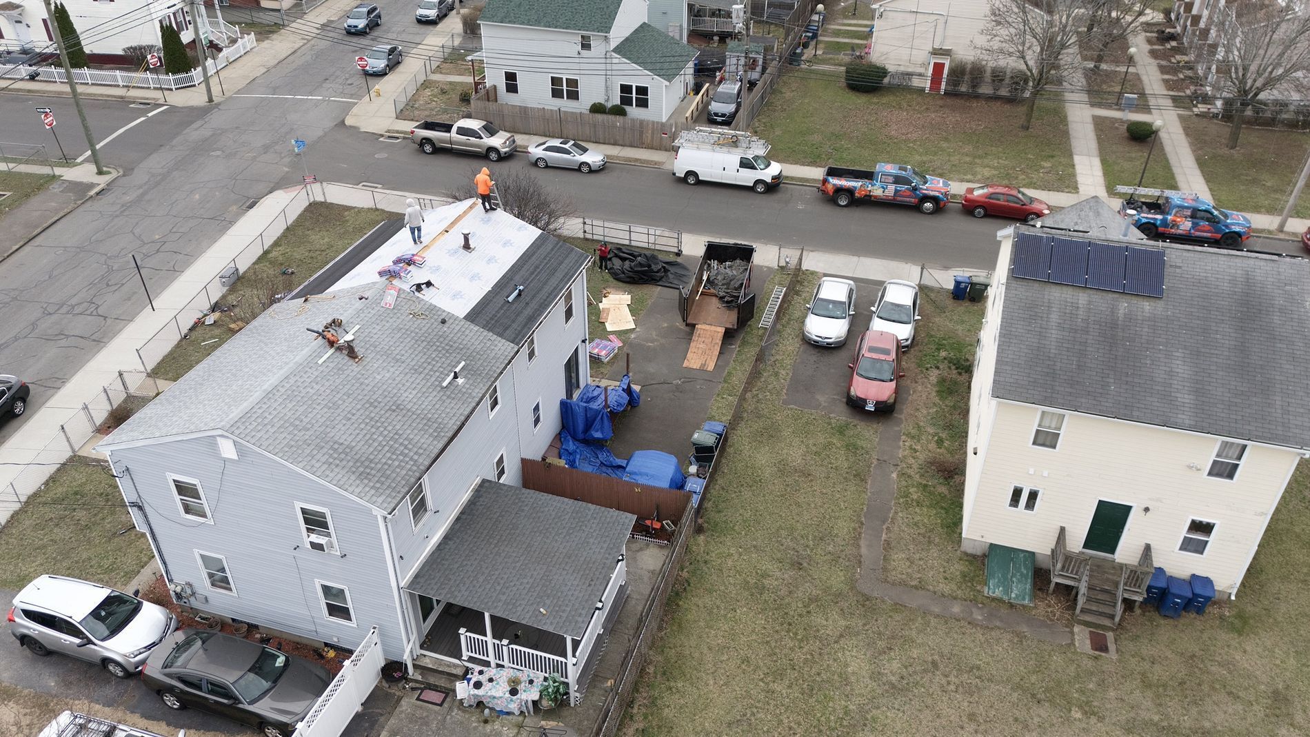Aerial view of two houses with a narrow driveway, parked cars, and a small backyard with a blue awning.