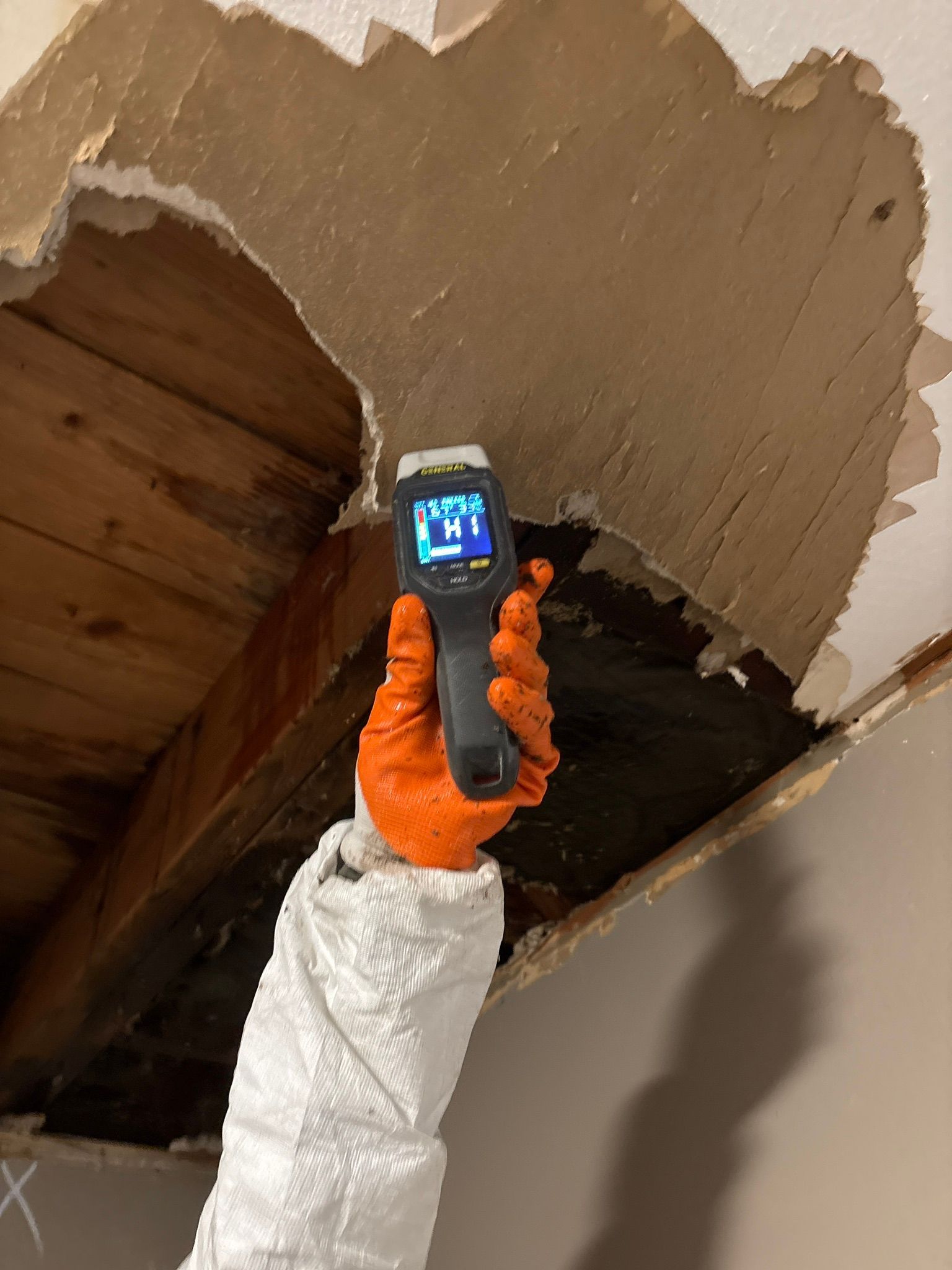 Gloved hand holds a thermal camera up to a damaged ceiling with exposed drywall and wooden beams