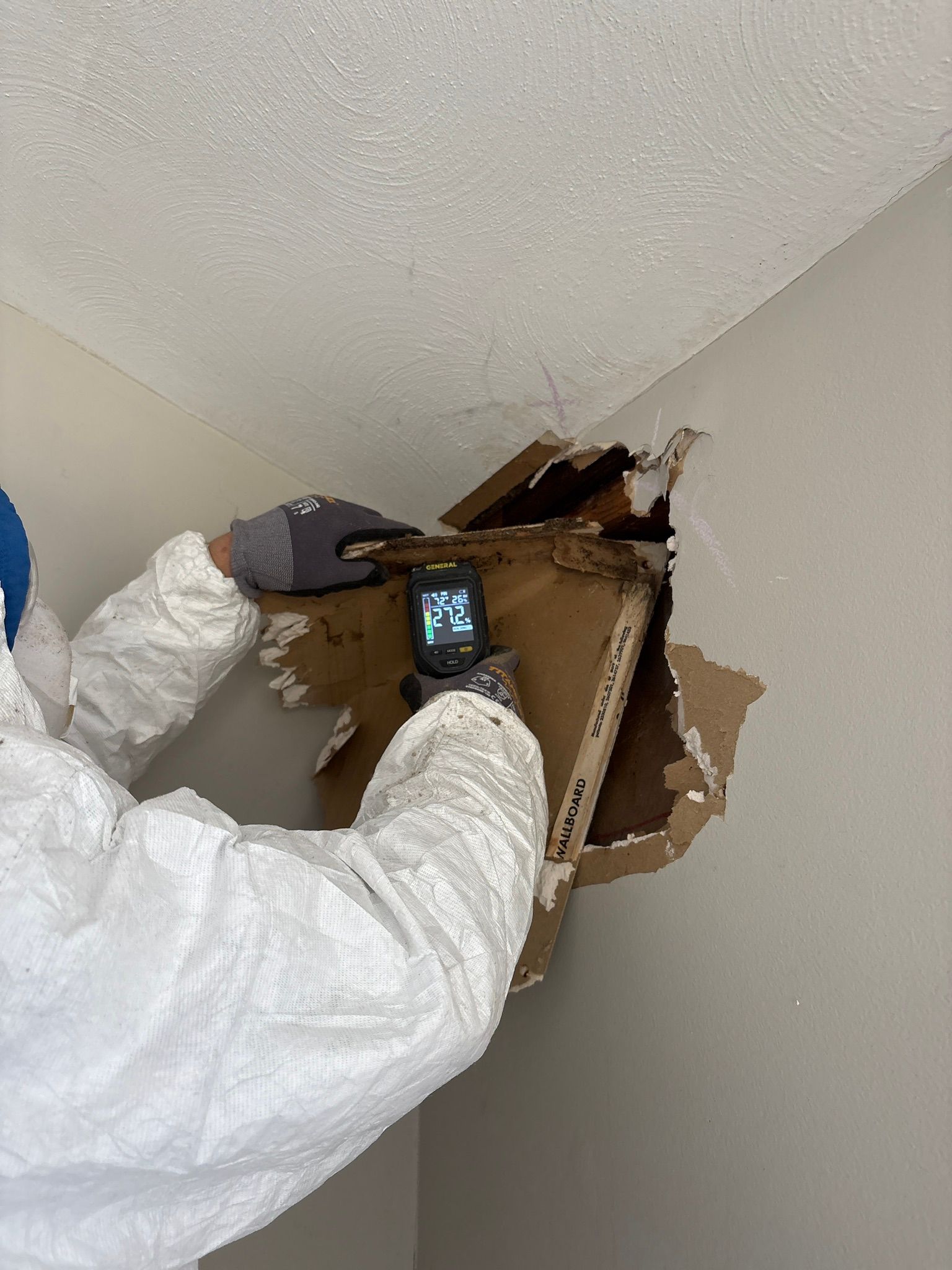 Person in white coveralls inspecting a large hole in a ceiling or wall with a phone or tool.