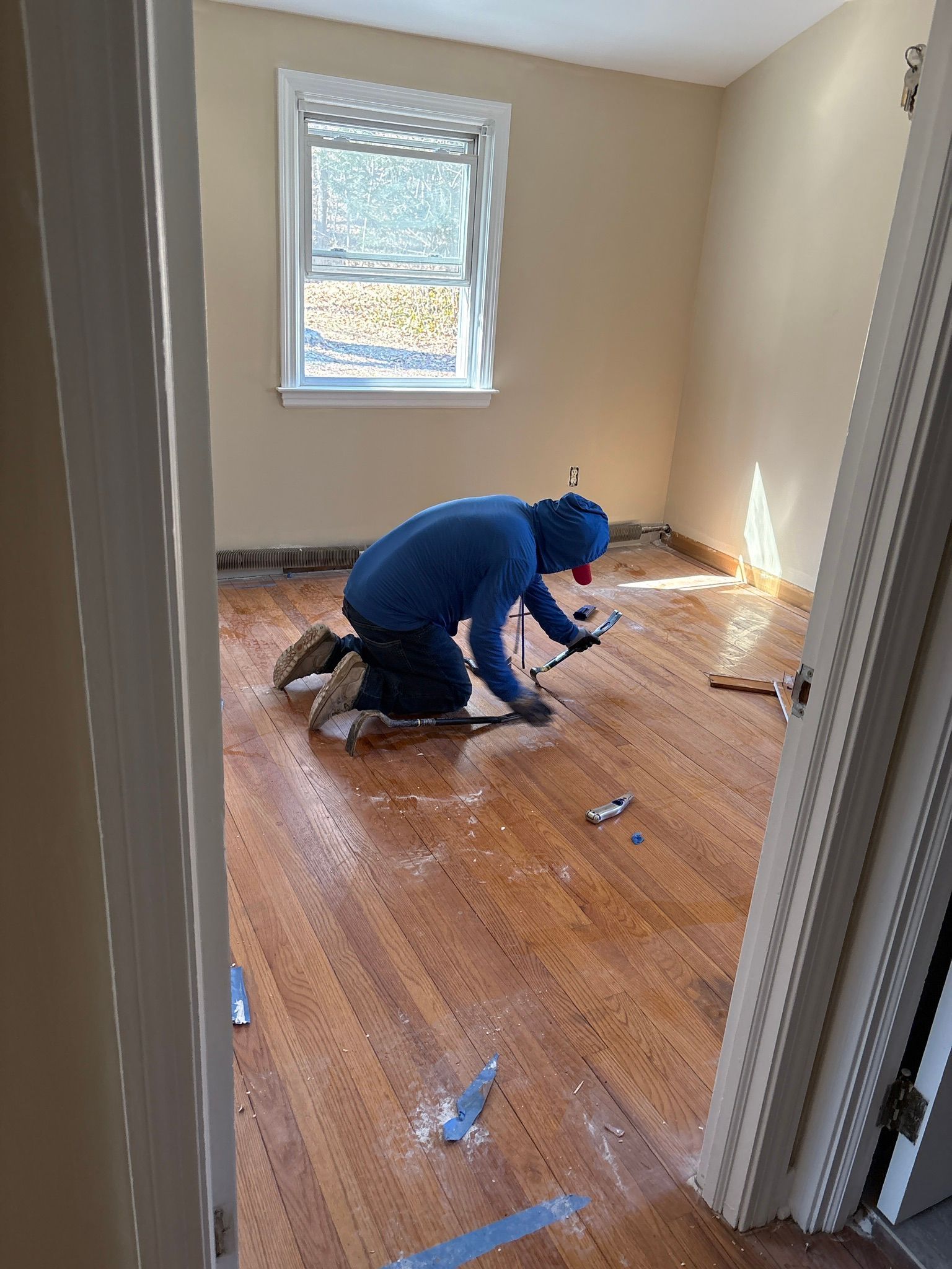 Person kneeling on hardwood floor, cleaning debris in a small beige room near a window.