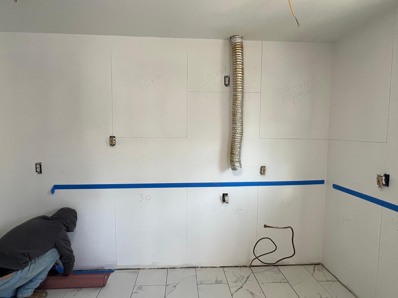 Worker taping a white room during renovation, with blue painter’s tape and exposed vent ducting.