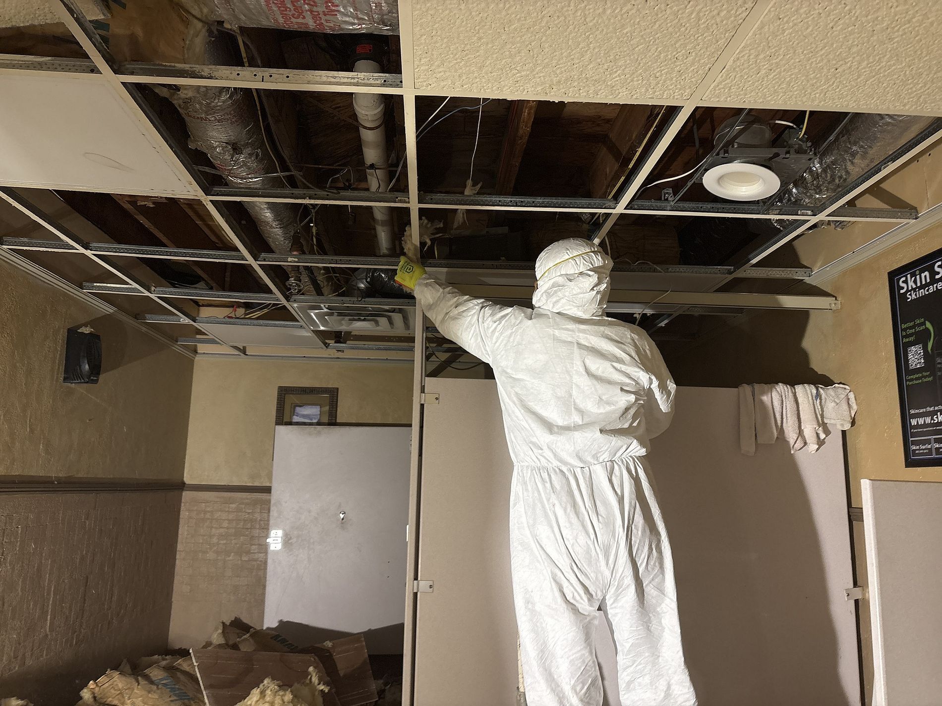 Worker in white suit inspecting a damaged ceiling with exposed insulation in a room under repair