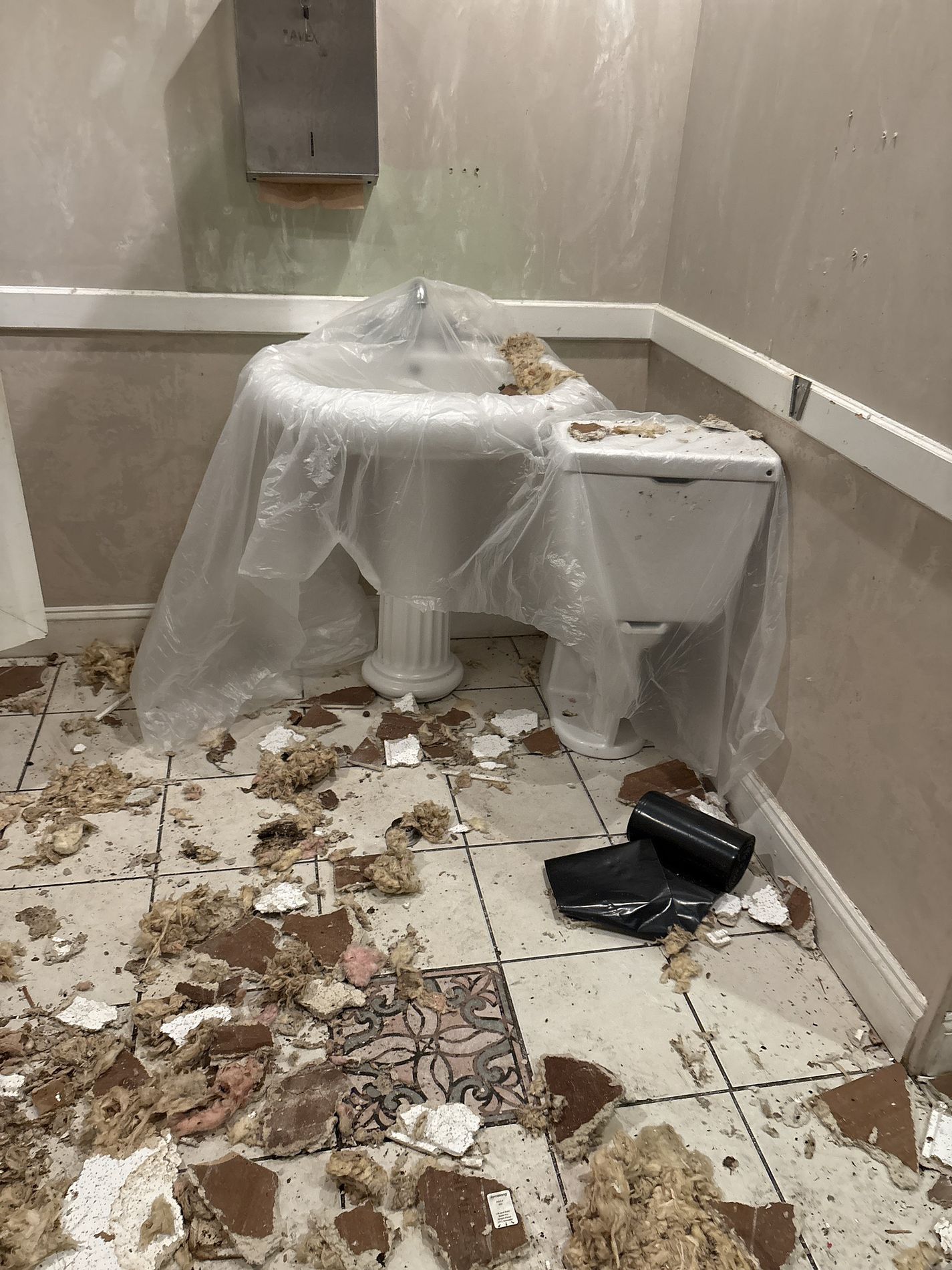 Damaged public restroom stall with toilet draped in plastic and debris scattered across the floor.