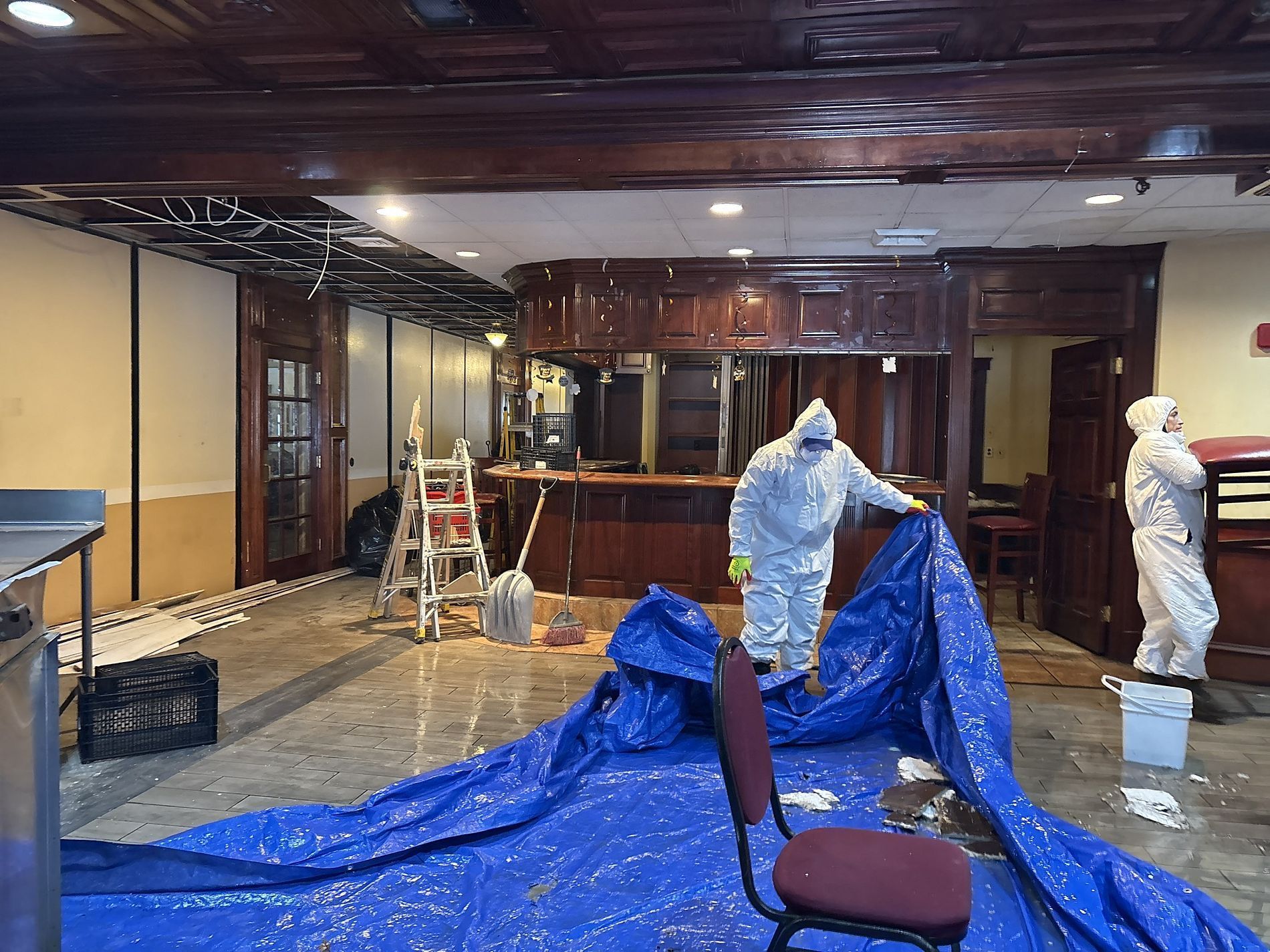 Workers in protective suits clean a flooded room with blue tarps and cleaning supplies.