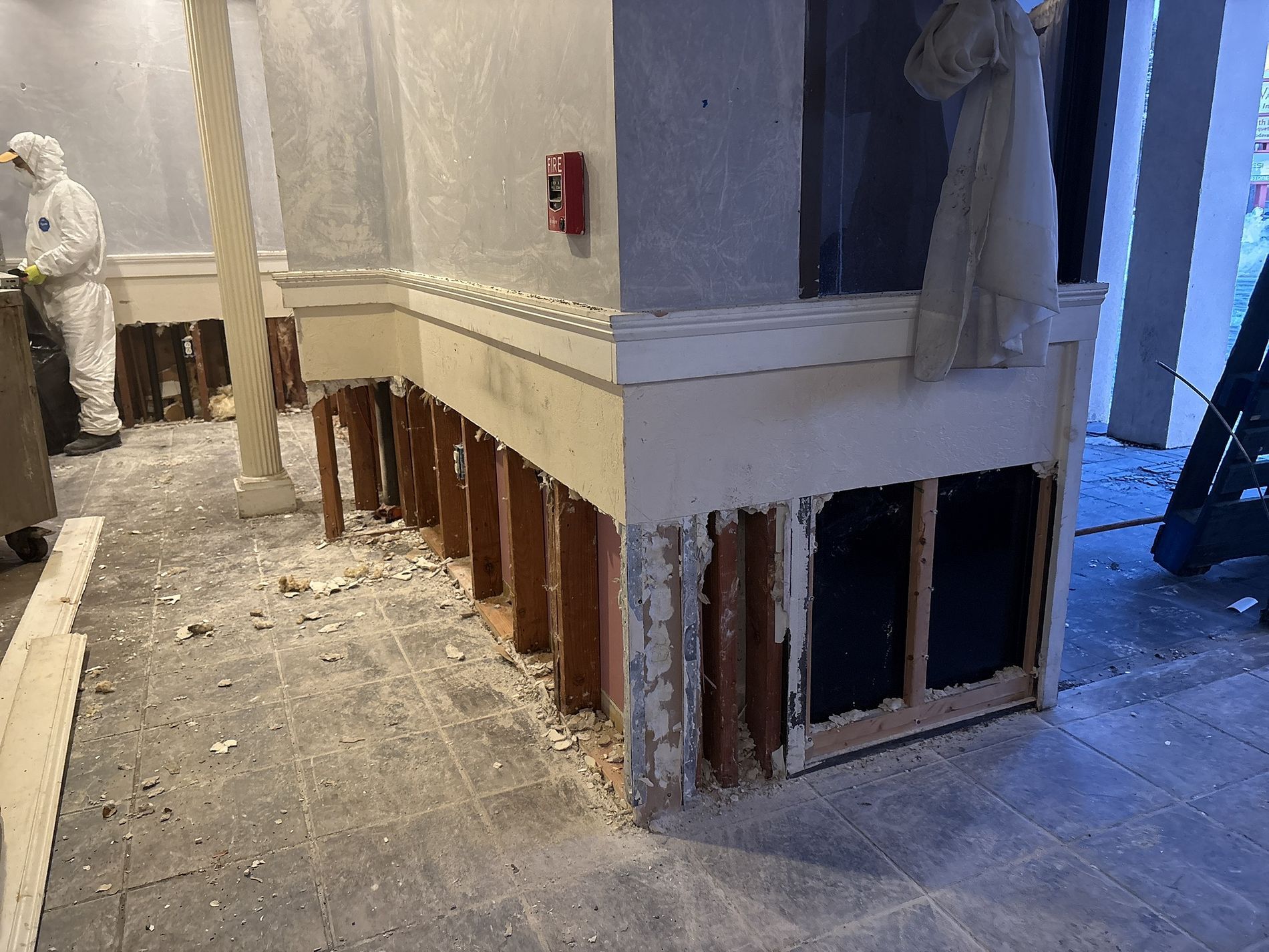 Damaged interior wall with exposed studs and debris along a hallway corner