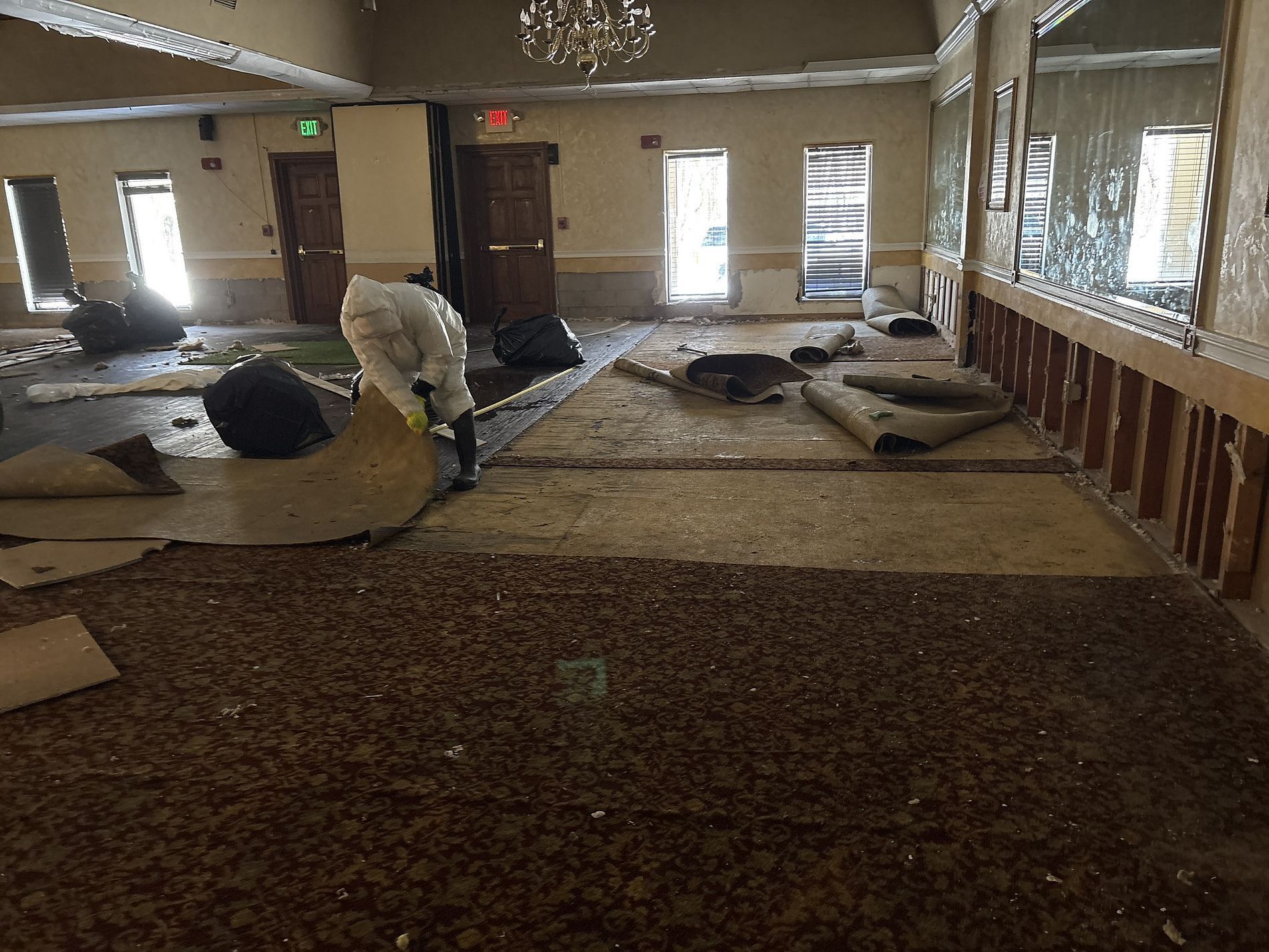 Indoor room with scattered mattresses and debris, a person in protective gear cleaning the floor.