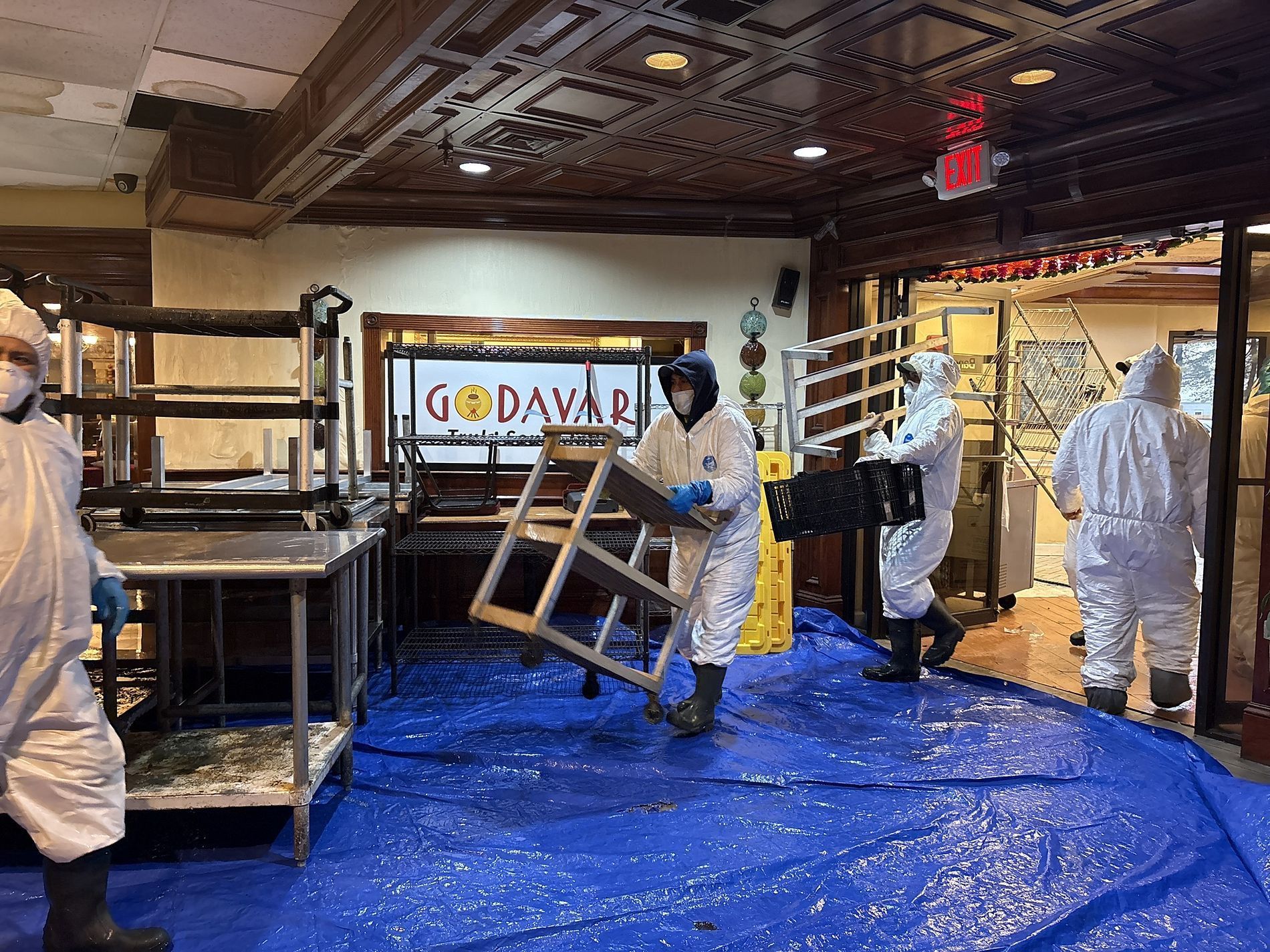Workers in white protective suits inside a flooded building, moving equipment over a blue tarp.