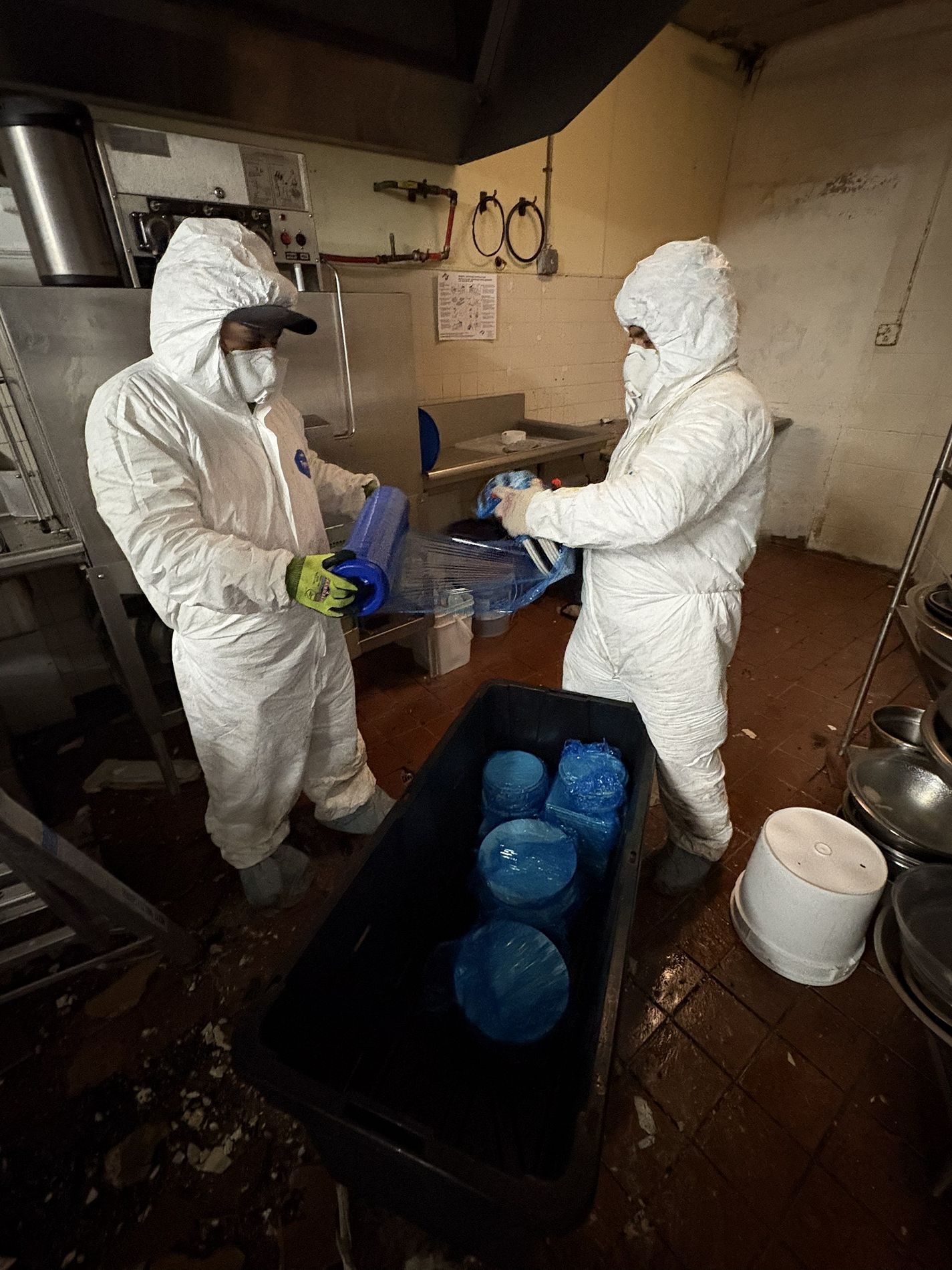 Two workers in white protective suits handle blue items in a dim, cluttered workshop.