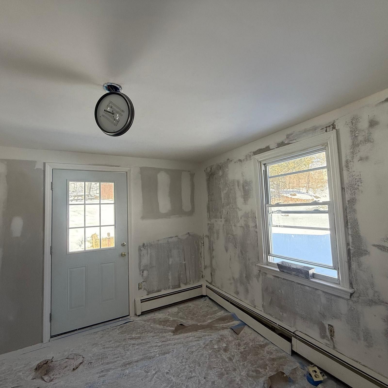 Small unfinished room with patched walls, two windows, a door, and a bare floor under renovation.