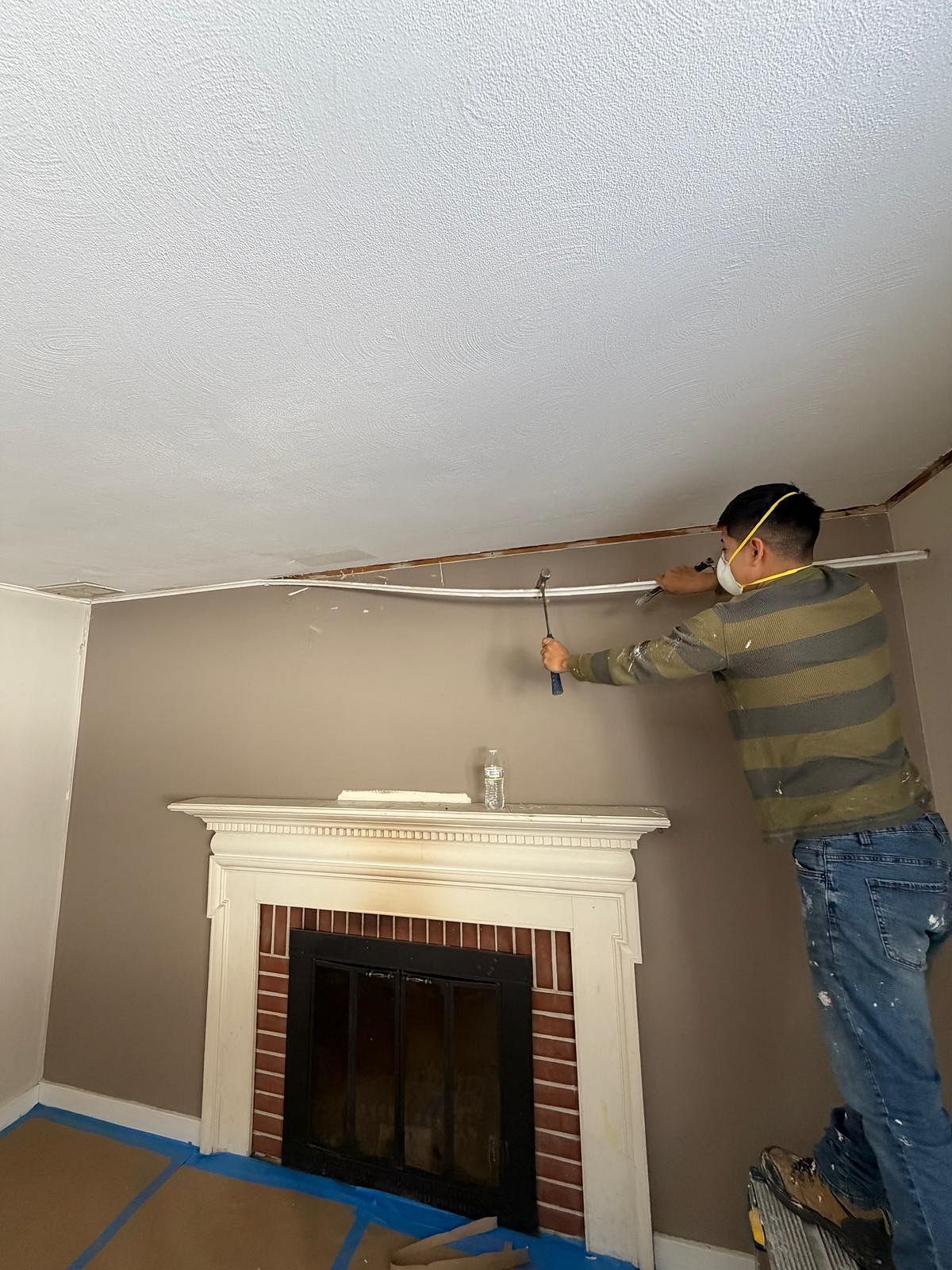 Worker installing trim above a fireplace in a room with taped-off floor and beige walls