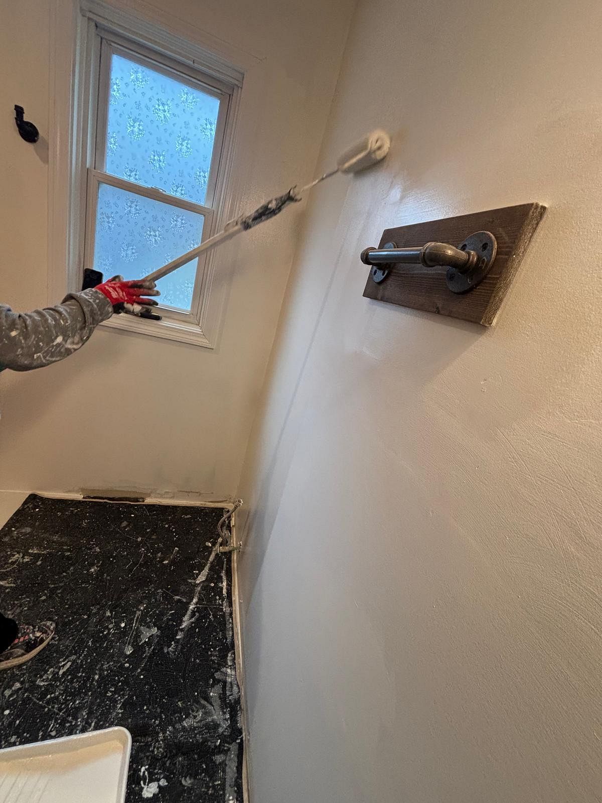 Person applying paint to a small white room with a roller and black floor protection