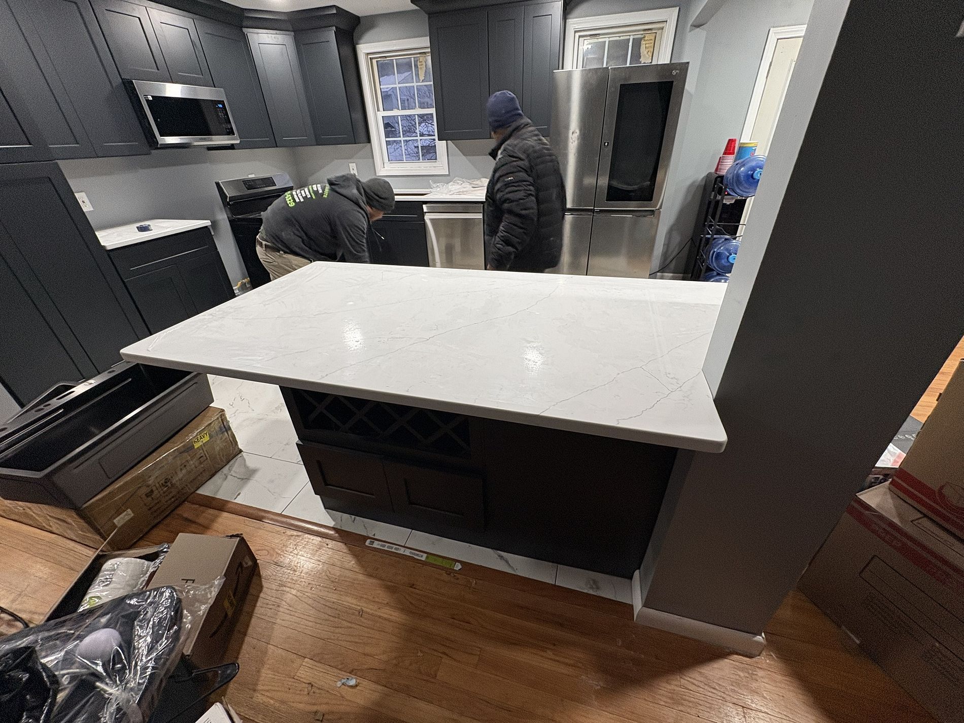 Kitchen under renovation with two workers behind a white island countertop and gray cabinetry.