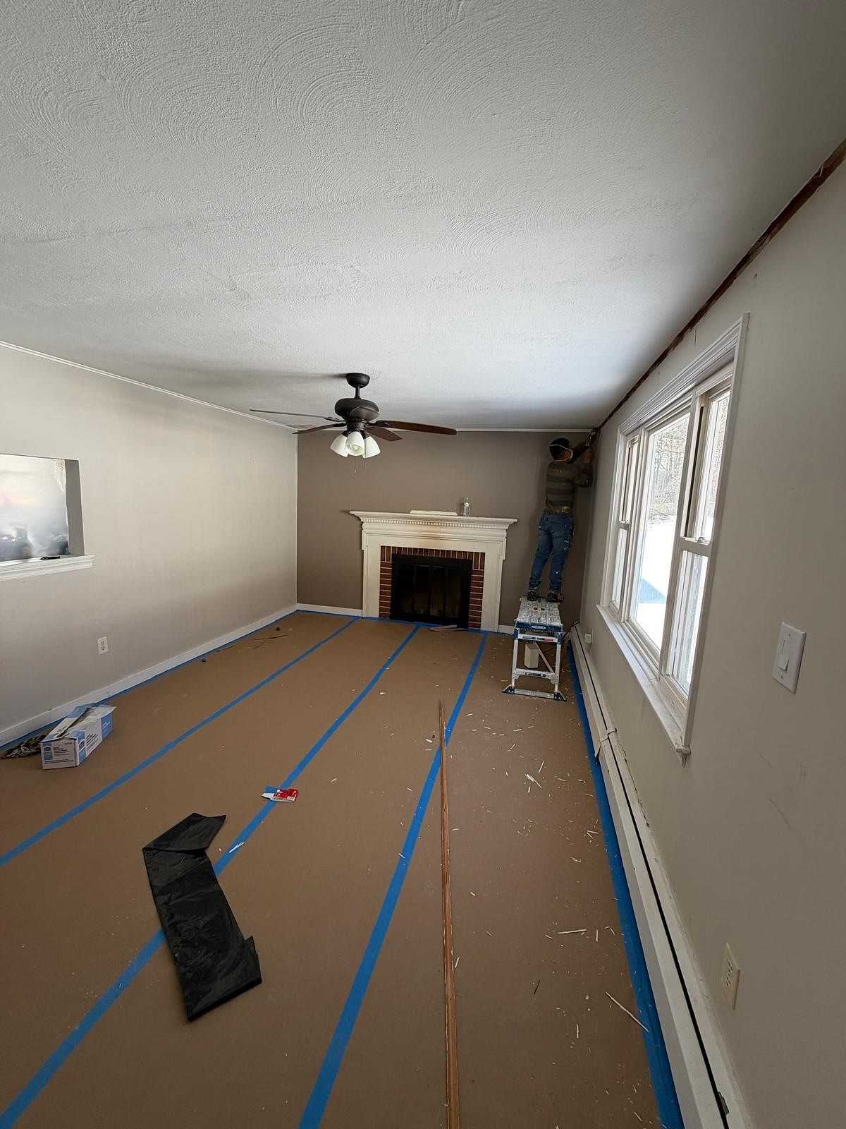 Living room under renovation with taped floor, fireplace, windows, ceiling fan, and ladders in the corner