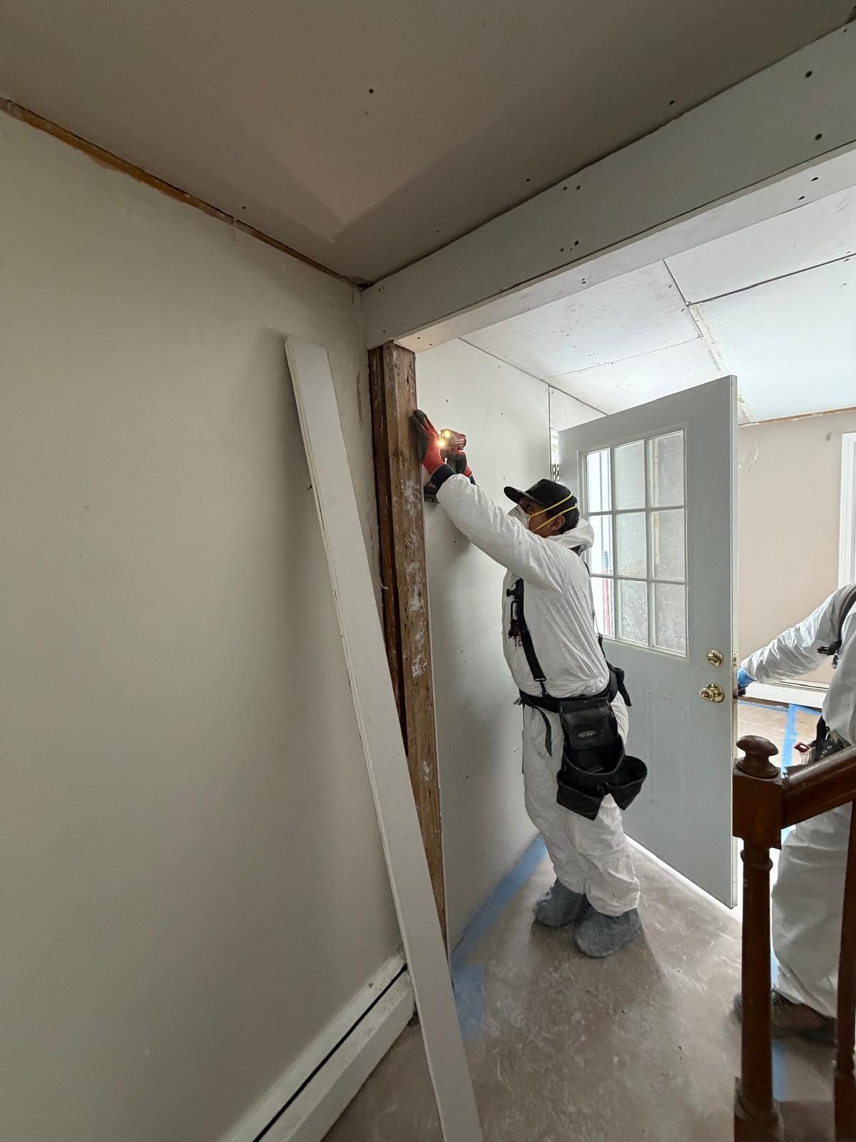 Worker sanding a wall corner in a bright room with plastic sheeting and a ladder nearby