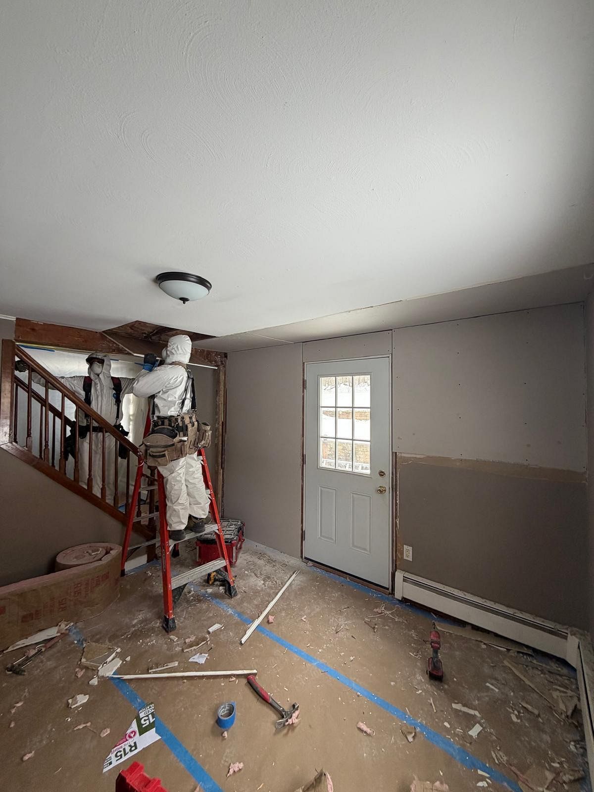 Partially renovated room with debris on the floor, staircase on the left, and a white door on the back wall
