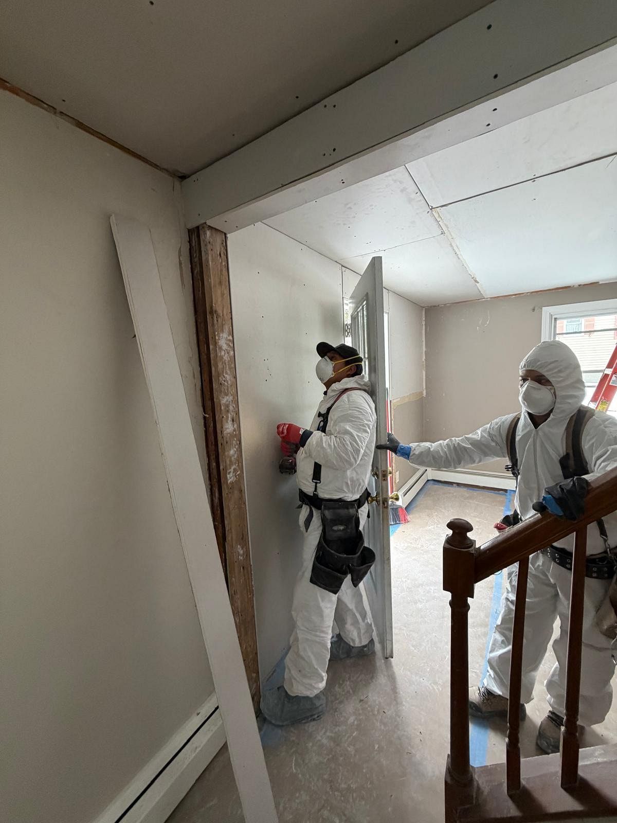 Two workers in white protective suits inspect a damaged interior hallway near exposed framing.