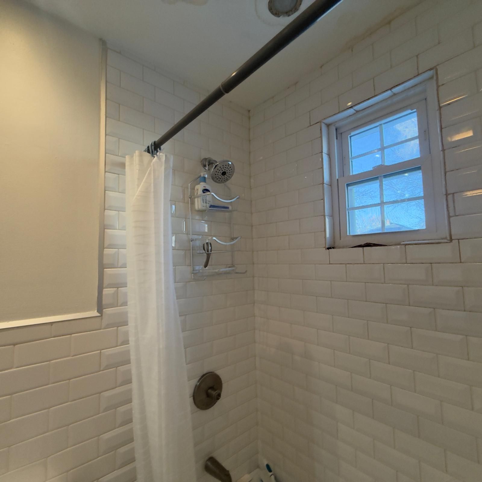 Small tiled shower with shower curtain, showerhead, and a window above the tub.