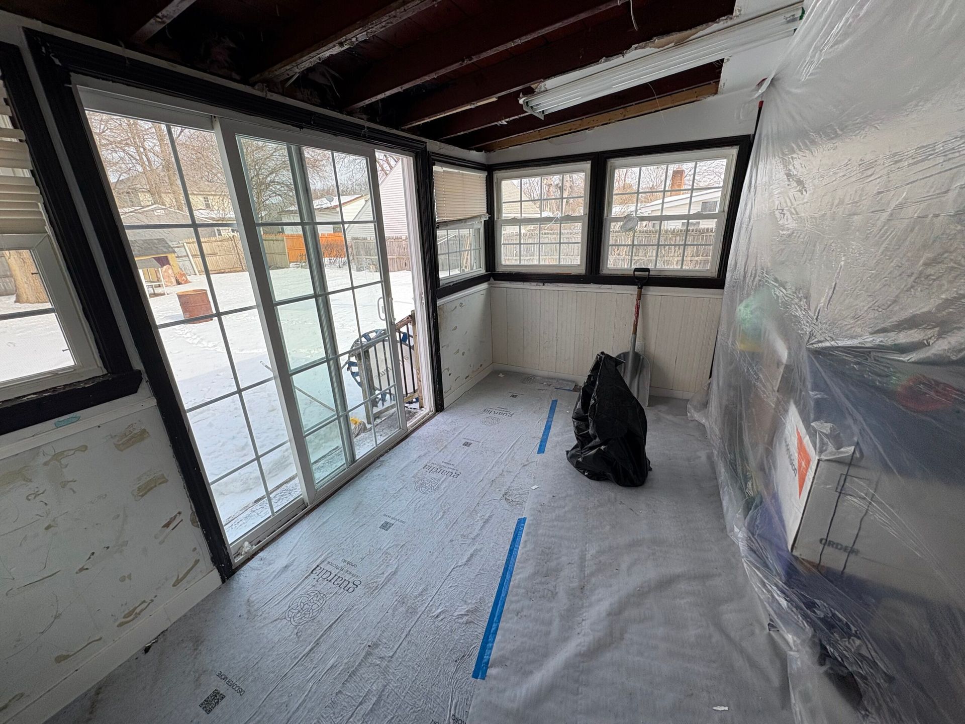Small room under renovation with plastic-covered floor, large windows, and a black backpack near the wall.