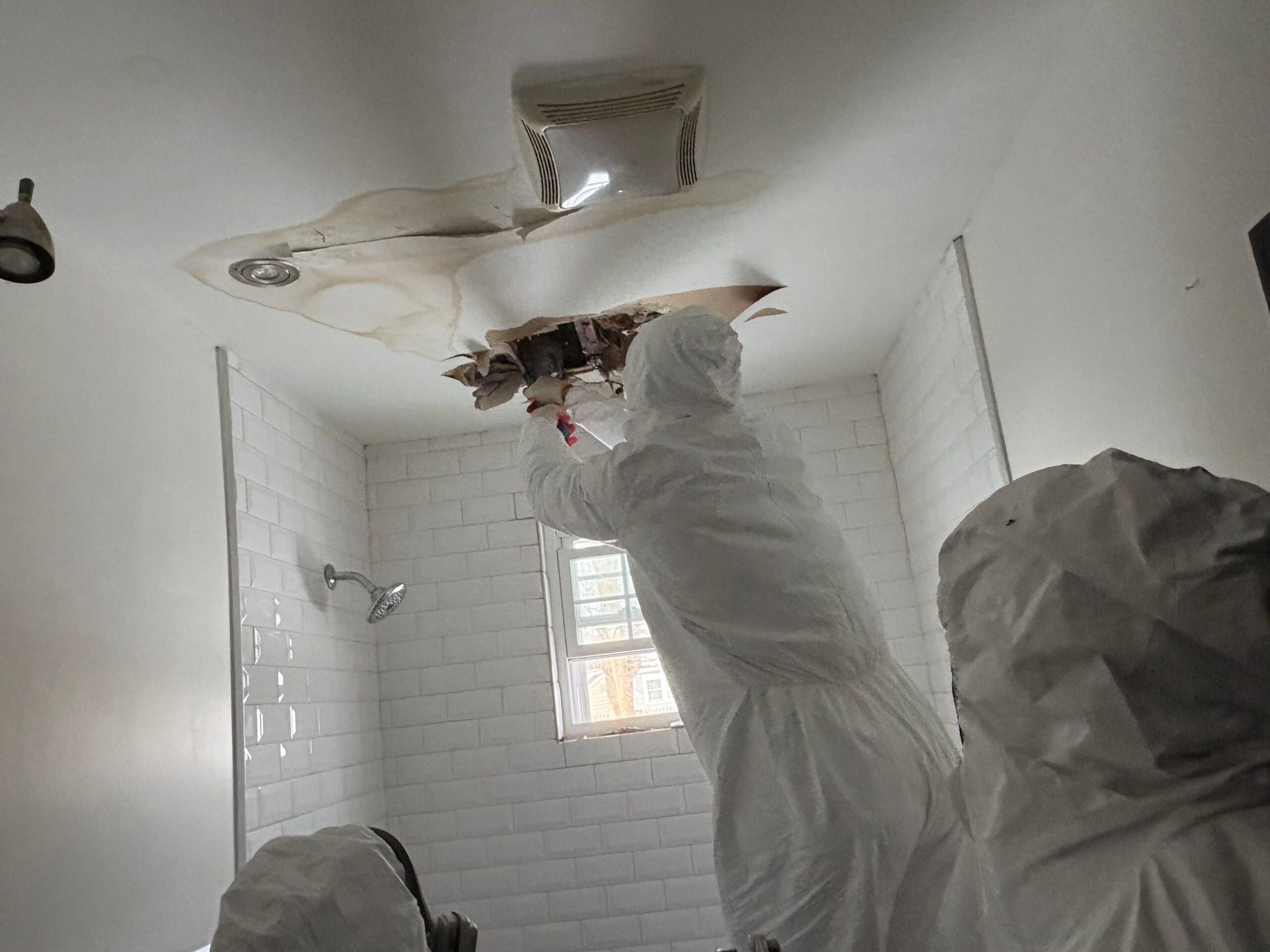 Worker in white coveralls repairing a damaged ceiling in a tiled bathroom.