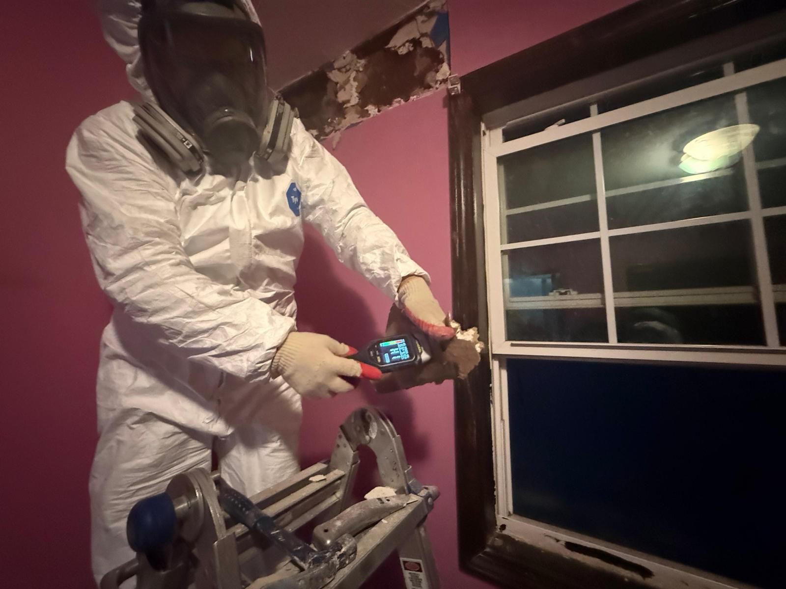 Person in a white suit and gas mask using a power tool beside an open window in a pink room