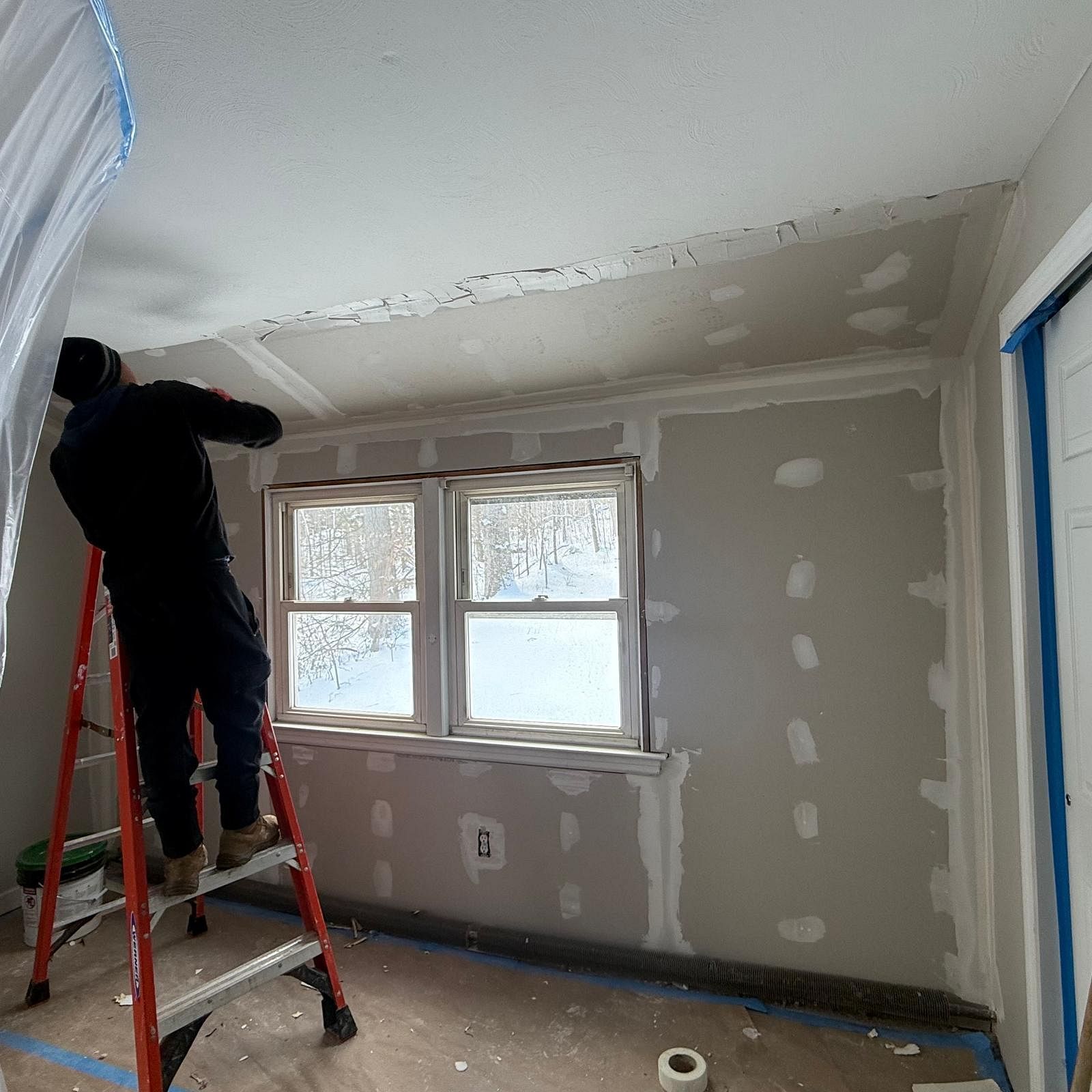 Worker on a ladder painting a gray bedroom wall with a window and blue painter’s tape.