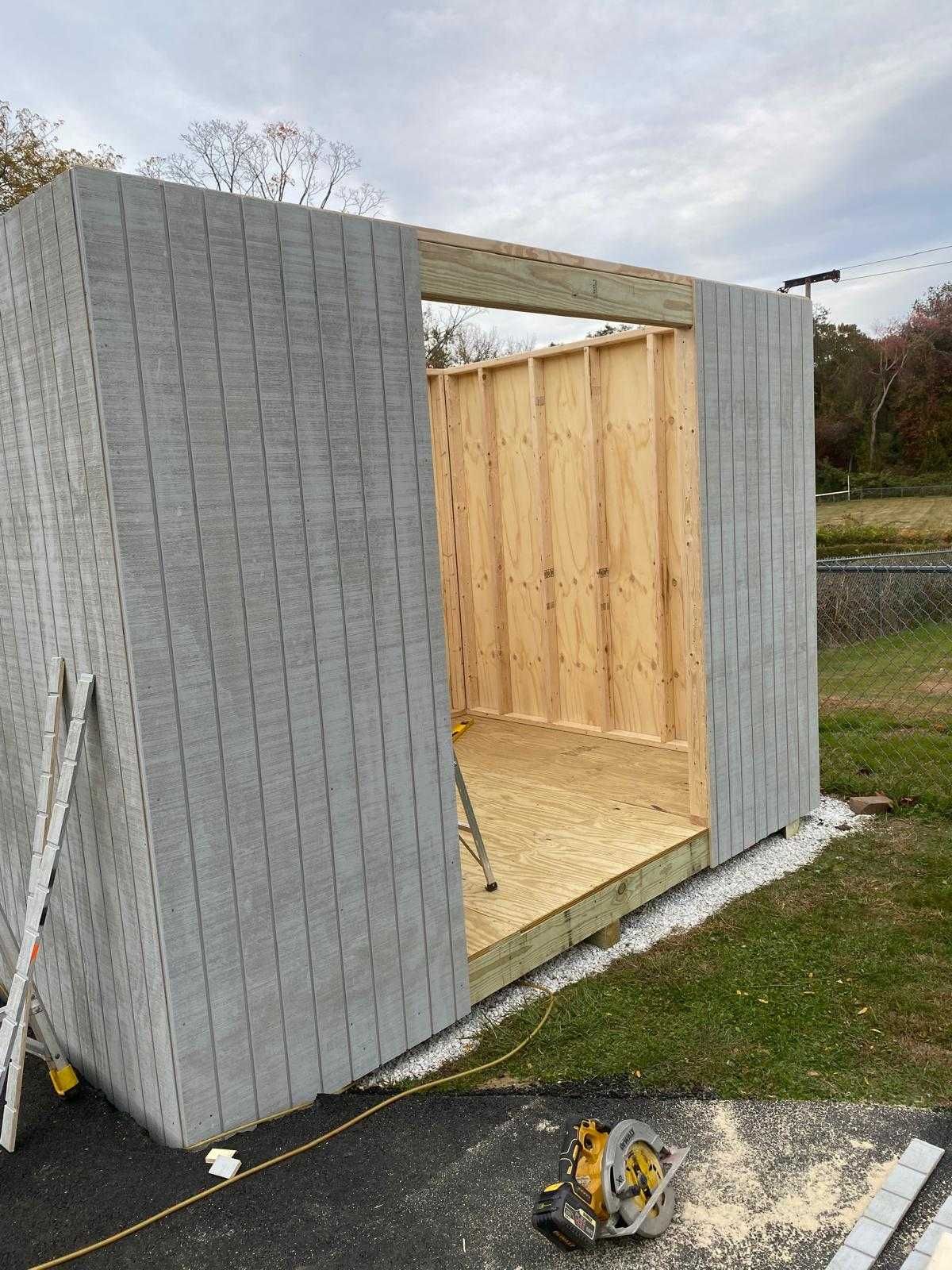 Partially built gray shed with an open doorway and exposed wooden interior, on a paved surface outdoors